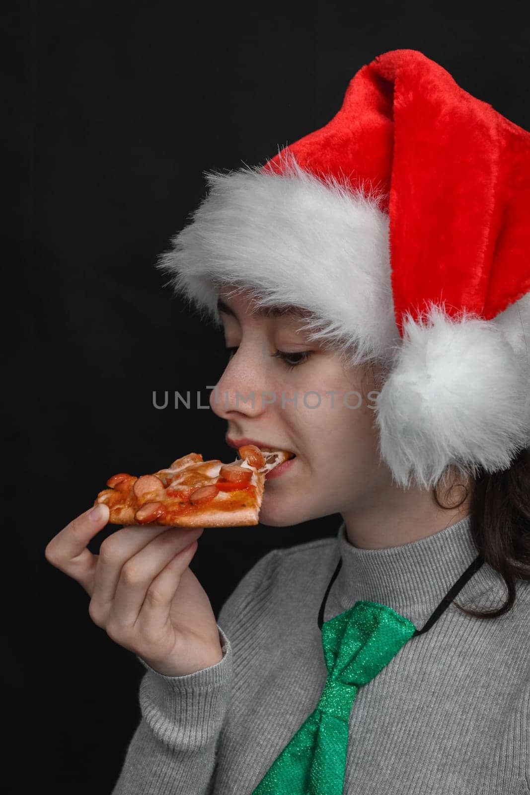 Happy caucasian teenage girl wearing a santa claus hat holds a slice of pizza in her hand and bites with her mouth with copy space for your text on a black background, close-up side view.