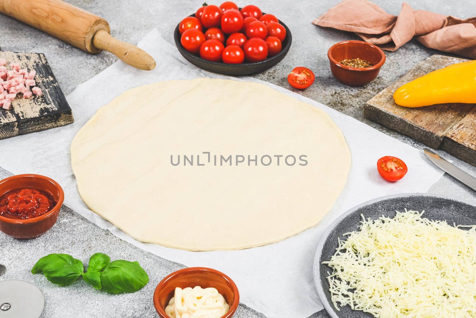 Pizza ingredients: cherry tomato, pepper, cheese, ham, basil with dough rolled out in a circle and a wooden chip lie on the table with copy space, side view close-up. Concept of cooking pizza.
