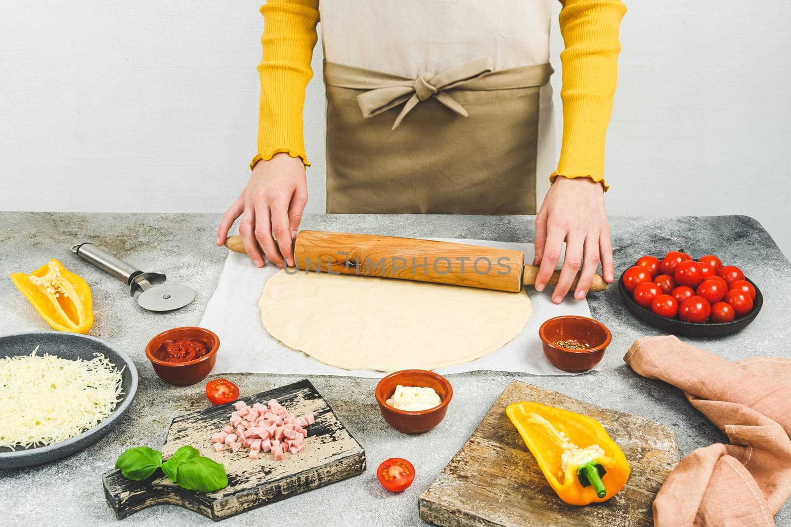 Hands of a caucasian teenager in an apron roll out pizza dough with a rolling pin on a table with ingredients, a knife and cutting boards, side view close-up.