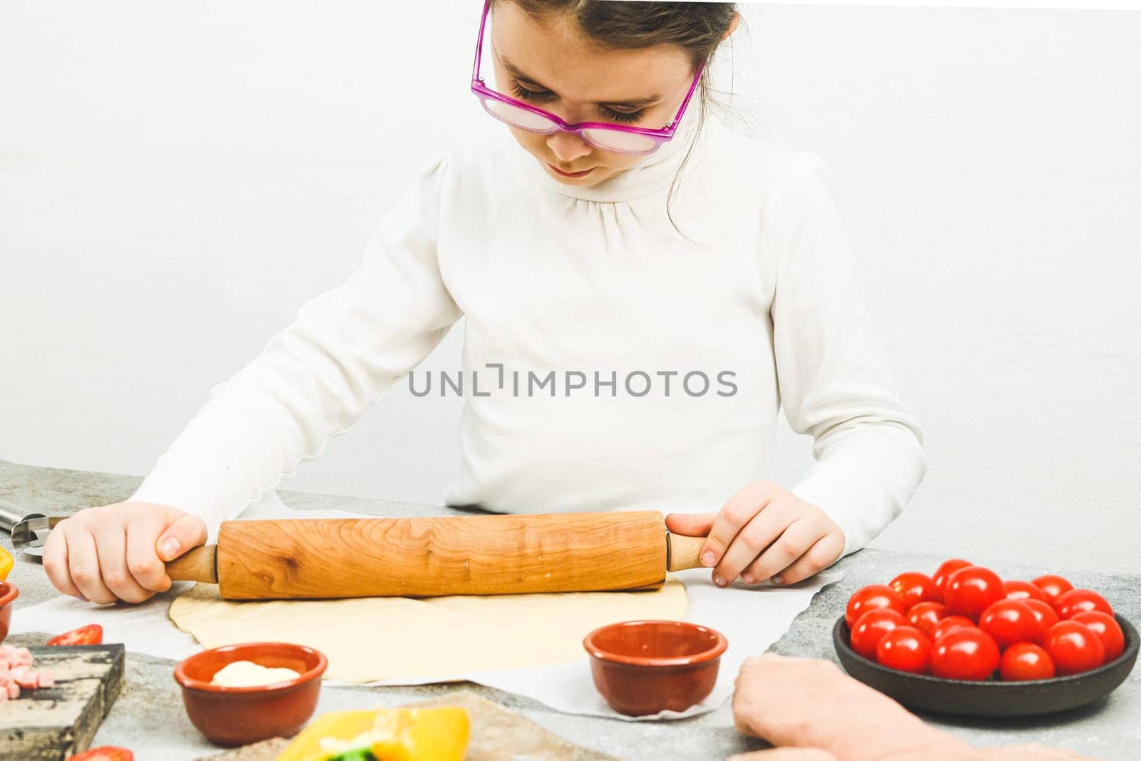 Caucasian girl in a white turtleneck with glasses rolls out pizza dough with a rolling pin on the table with ingredients, a knife and cutting boards, side view close-up. Concept of cooking pizza.