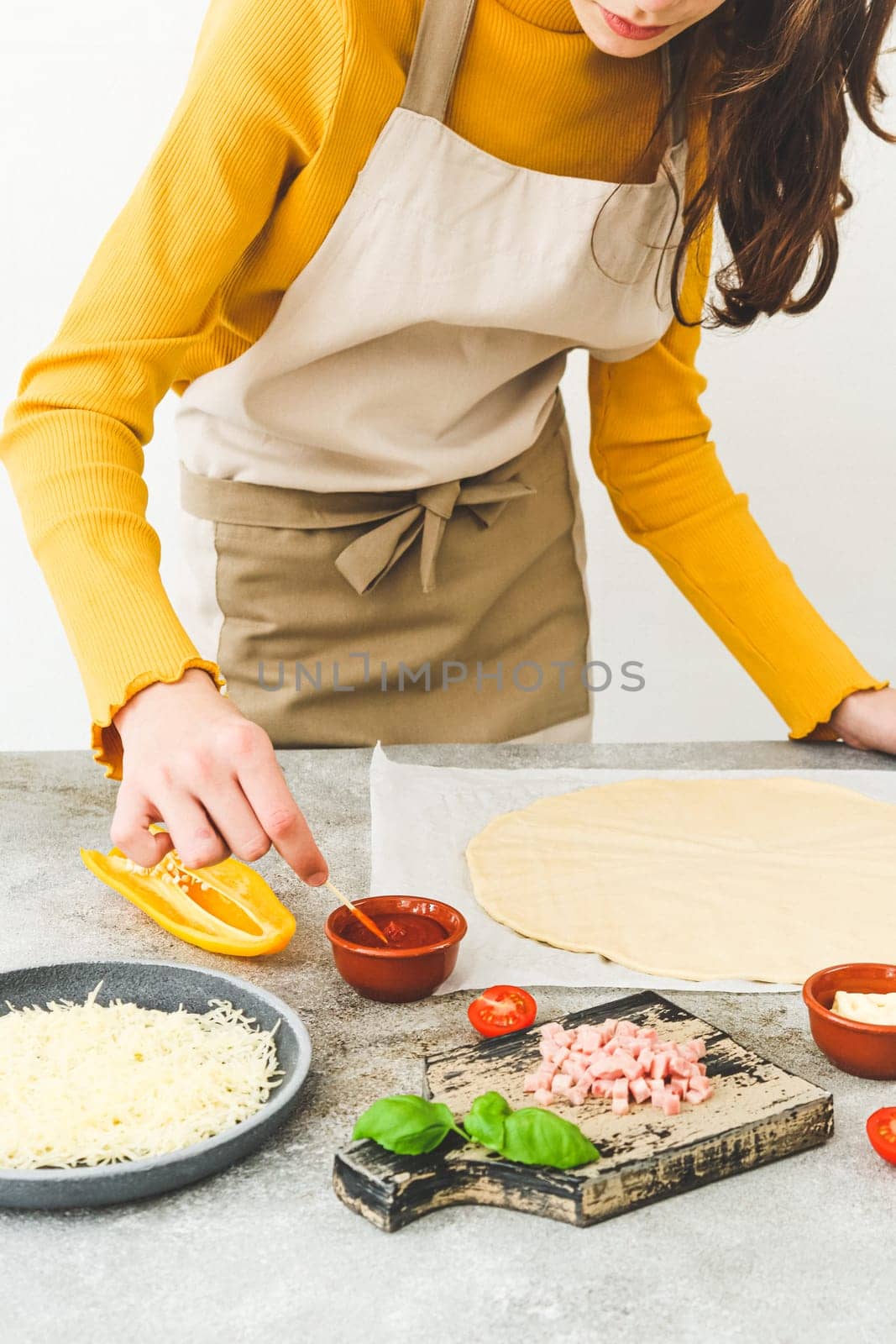 Caucasian teenage girl in an apron at the table with ingredients and dough mixes the sauce for making pizza with a toothpick,