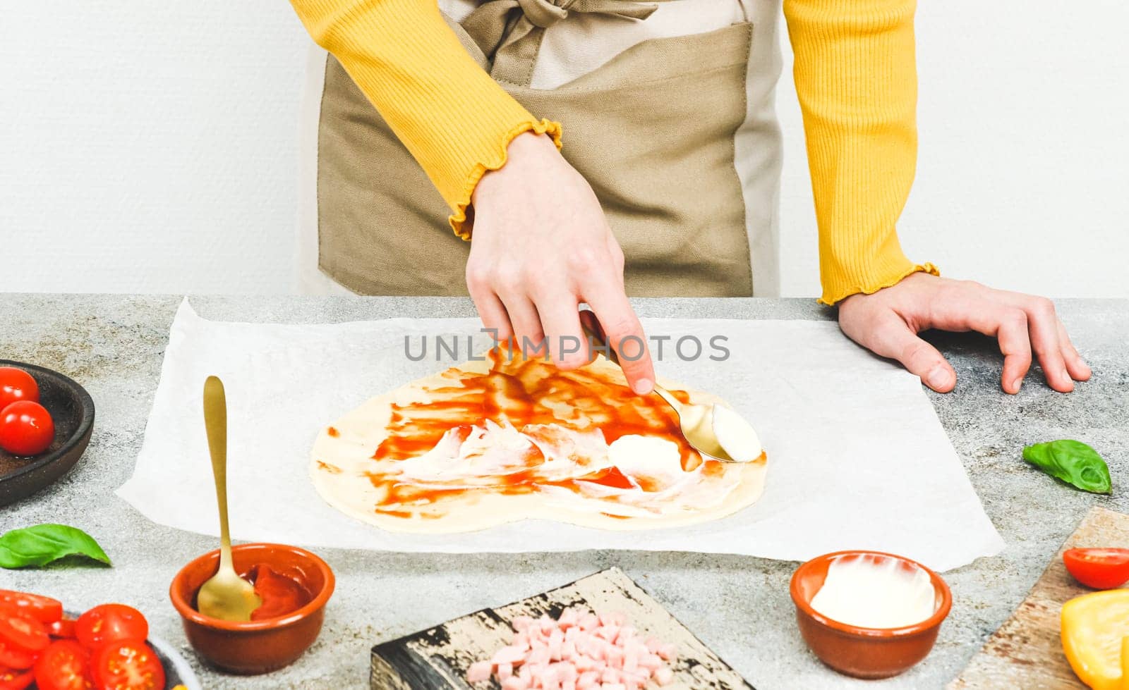 Hands of a Caucasian teenage girl smear a spoonful of pizza sauce on a heart-shaped dough on baking paper with wooden boards and pizza ingredients lie on a light gray table, side view close-up.