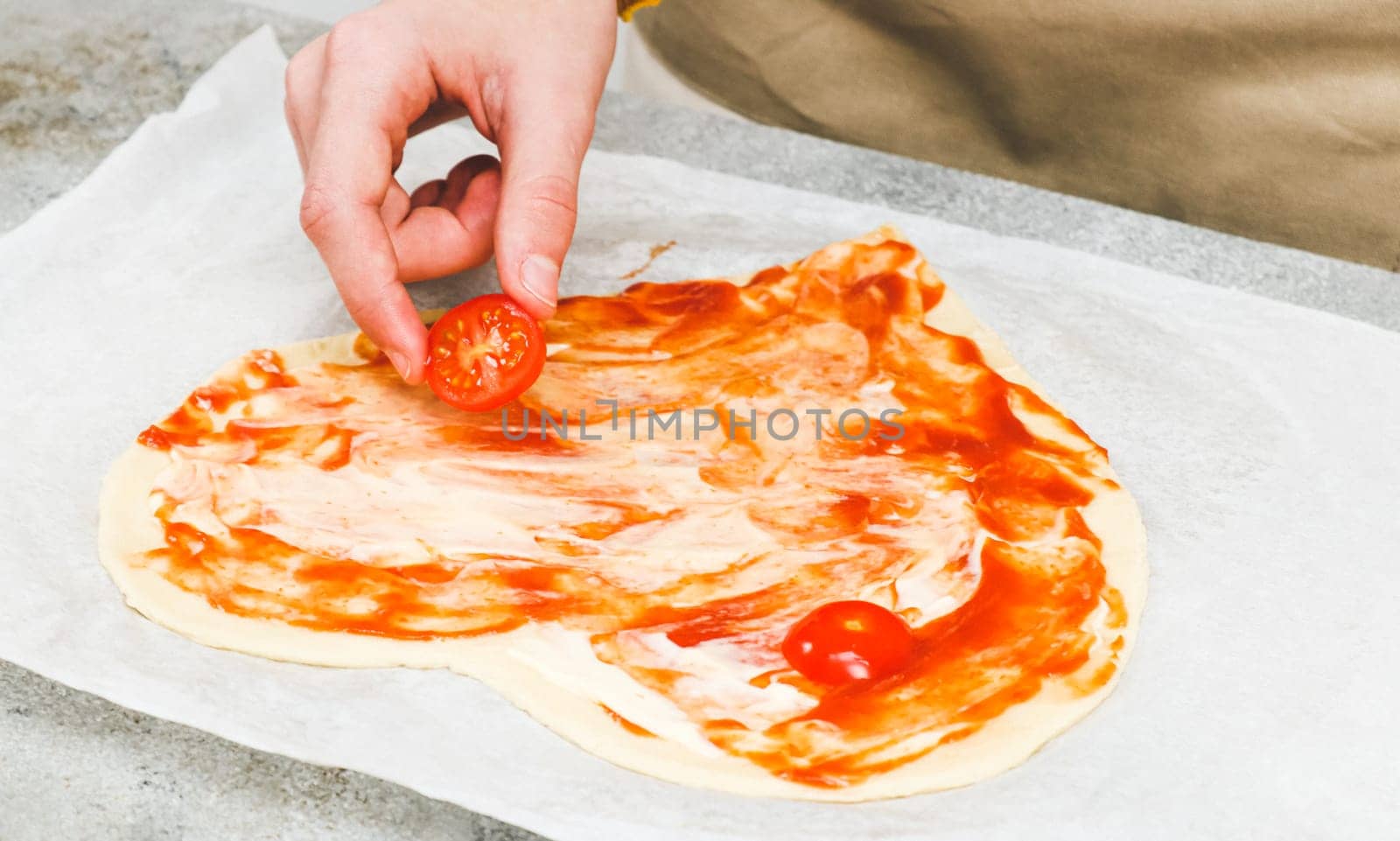 Hands of caucasian teenage girl lay down sliced cherry tomato on heart-shaped pizza dough for valentine's day with ingredients on the table, close-up side view. Valentine's day pizza making concept.