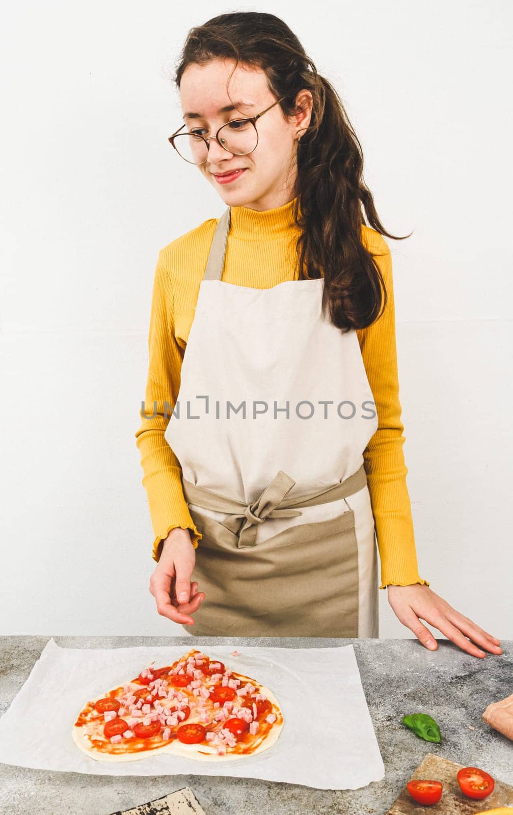 Caucasian teenage girl in an apron with a happy look looks to the side while standing at the table with raw pizza in the shape of a heart on parchment, side view close-up. Concept of cooking pizza.
