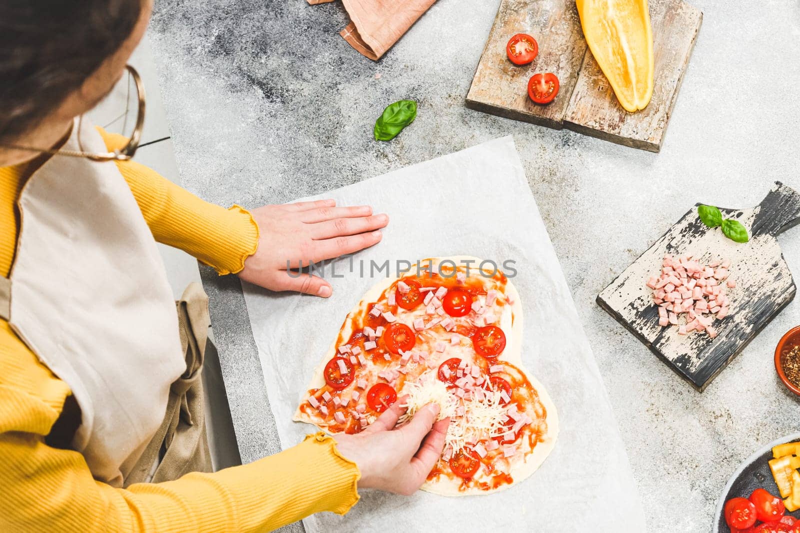 Caucasian teenage girl in an apron pouring grated cheese on pizza hearts for valentine's day with ingredients on the table, close-up side view. Concept of cooking pizza for valentine's day.