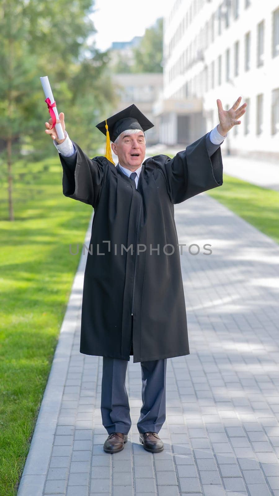 An elderly gray-haired man in a graduate robe spread his arms to the sides and holds a diploma outdoors. Vertical