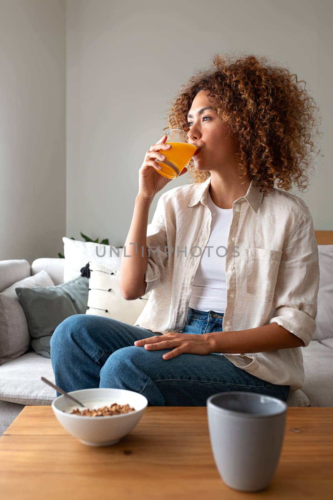 Vertical portrait of young multiracial woman drinking orange juice for breakfast sitting on sofa looking away pensive. by Hoverstock