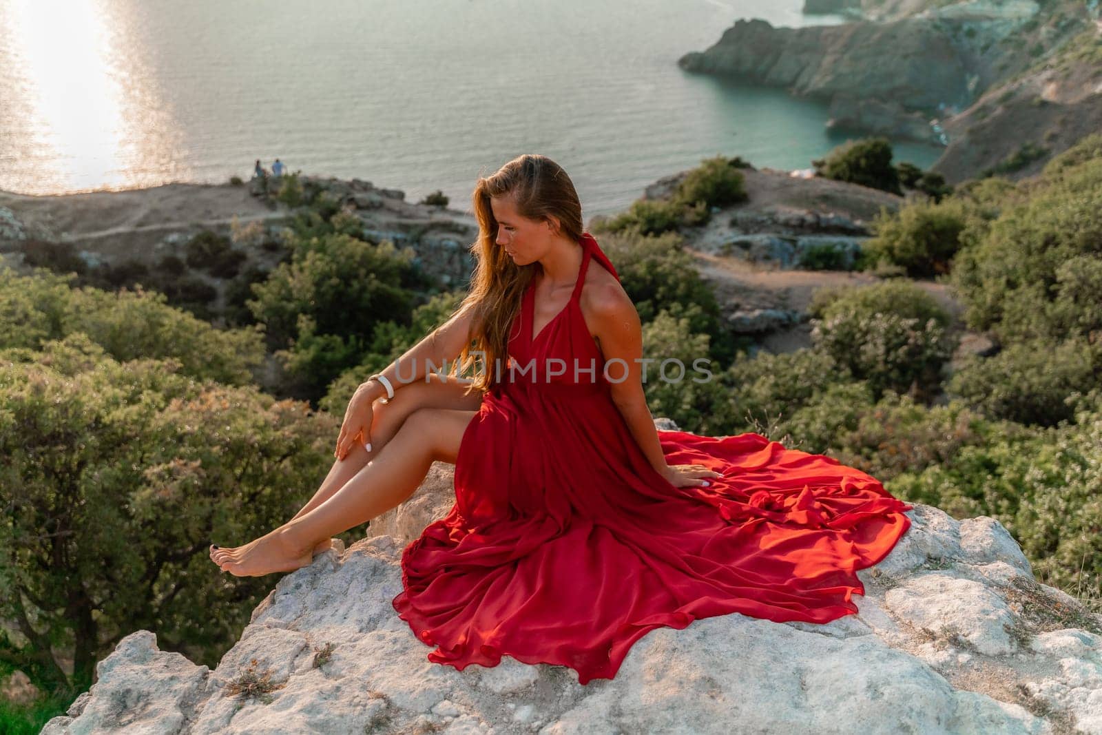 Woman sunset sea red dress, side view a happy beautiful sensual woman in a red long dress posing on a rock high above the sea on sunset. by Matiunina