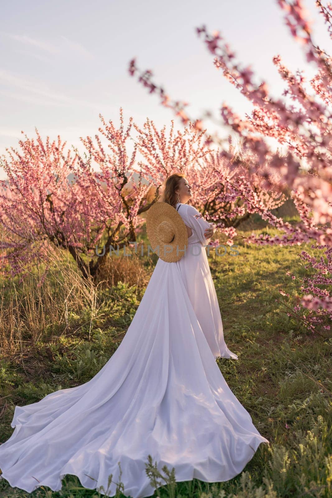 Woman blooming peach orchard. Against the backdrop of a picturesque peach orchard, a woman in a long white dress and hat enjoys a peaceful walk in the park, surrounded by the beauty of nature