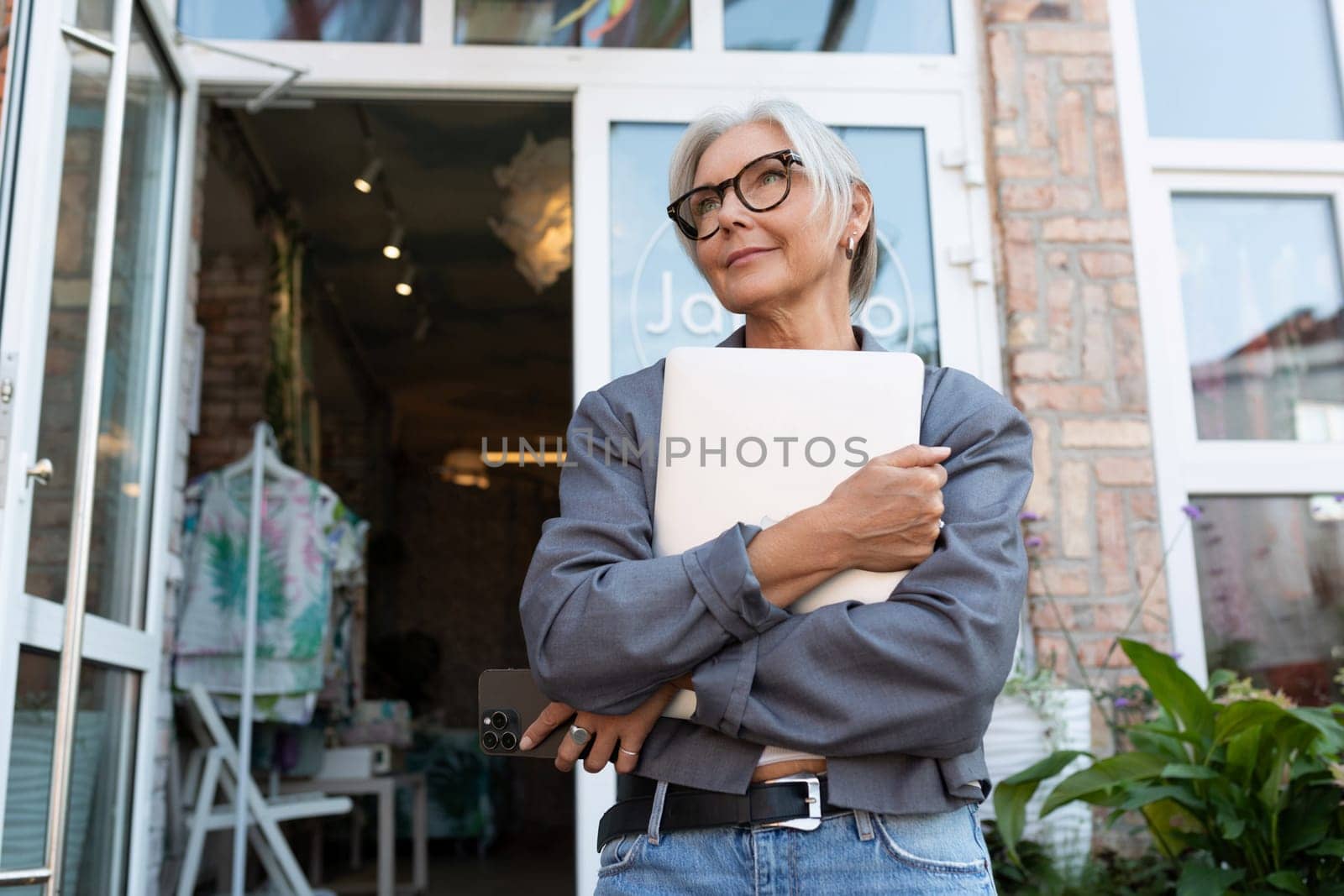 stylish mature woman with gray hair and glasses dressed in a gray jacket holds a laptop for work and business by TRMK