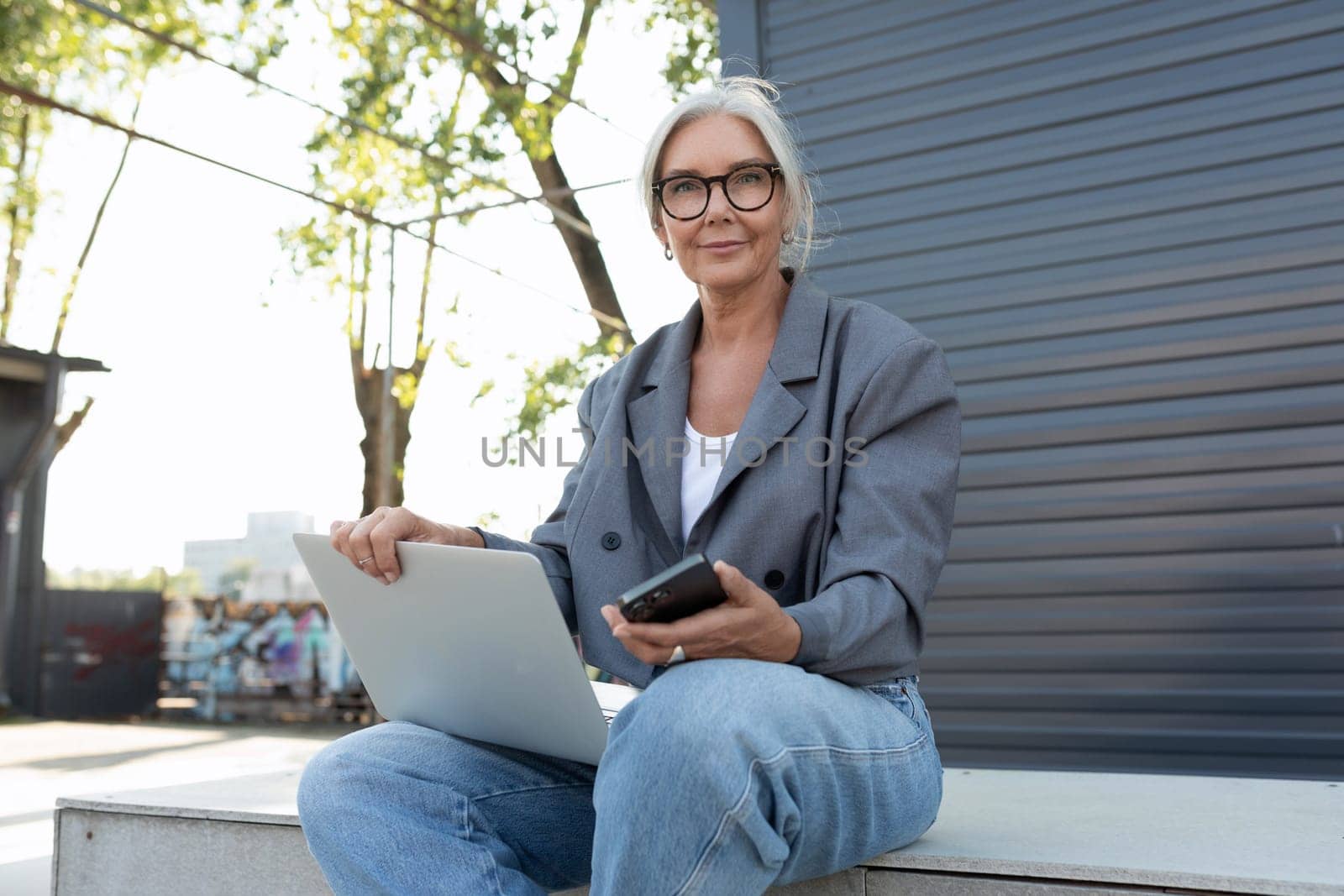 a gray-haired mature pretty business woman dressed in a stylish gray jacket and jeans sat down on a bench with a laptop by TRMK