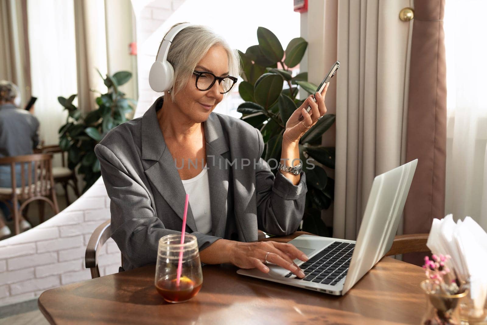 charming stylish mature business woman with glasses dressed in a gray jacket spends time in a cafe sitting with a laptop.