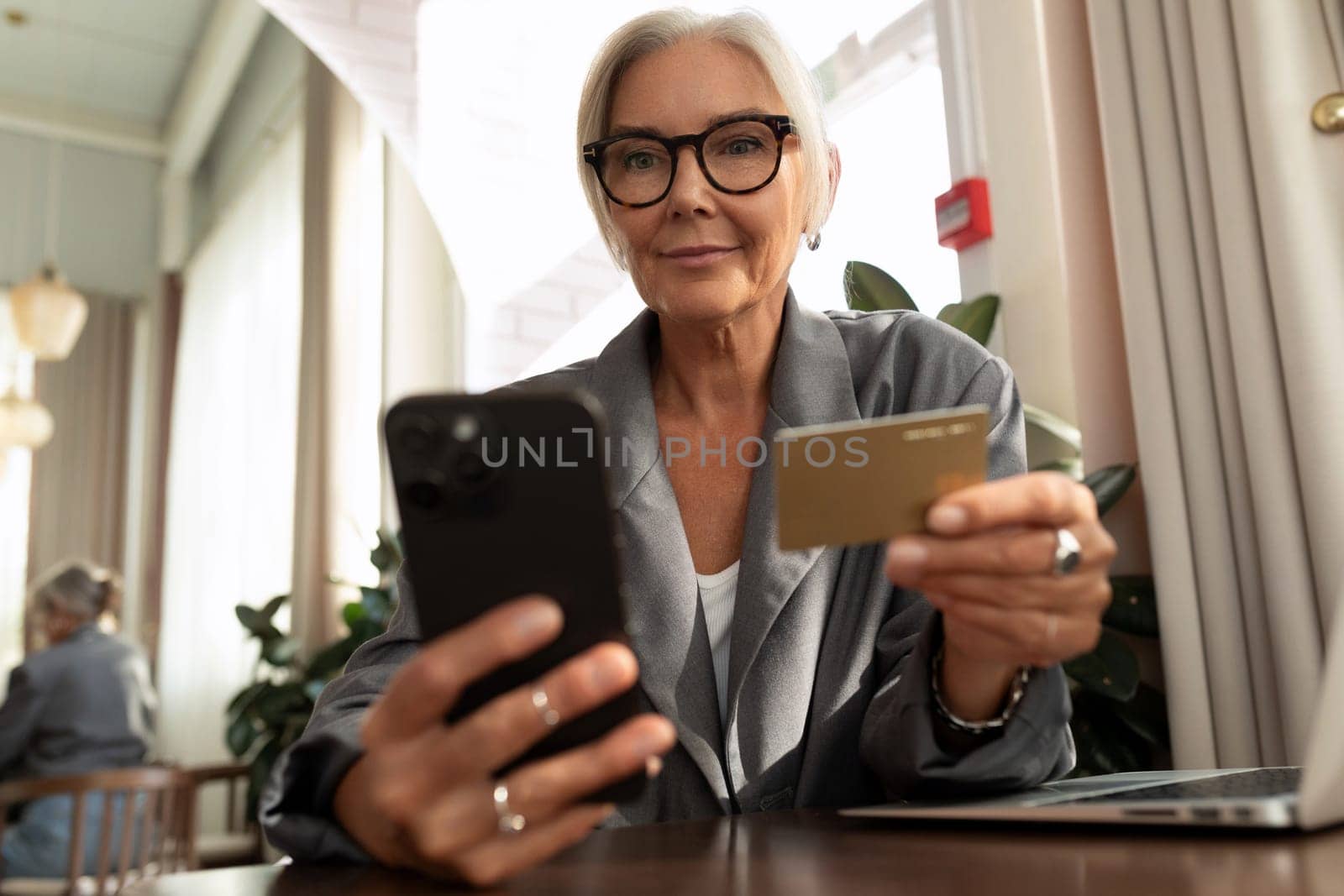 gray-haired mature pretty business woman dressed in a stylish gray jacket pays the bill in a cafe using a smartphone and a plastic card.