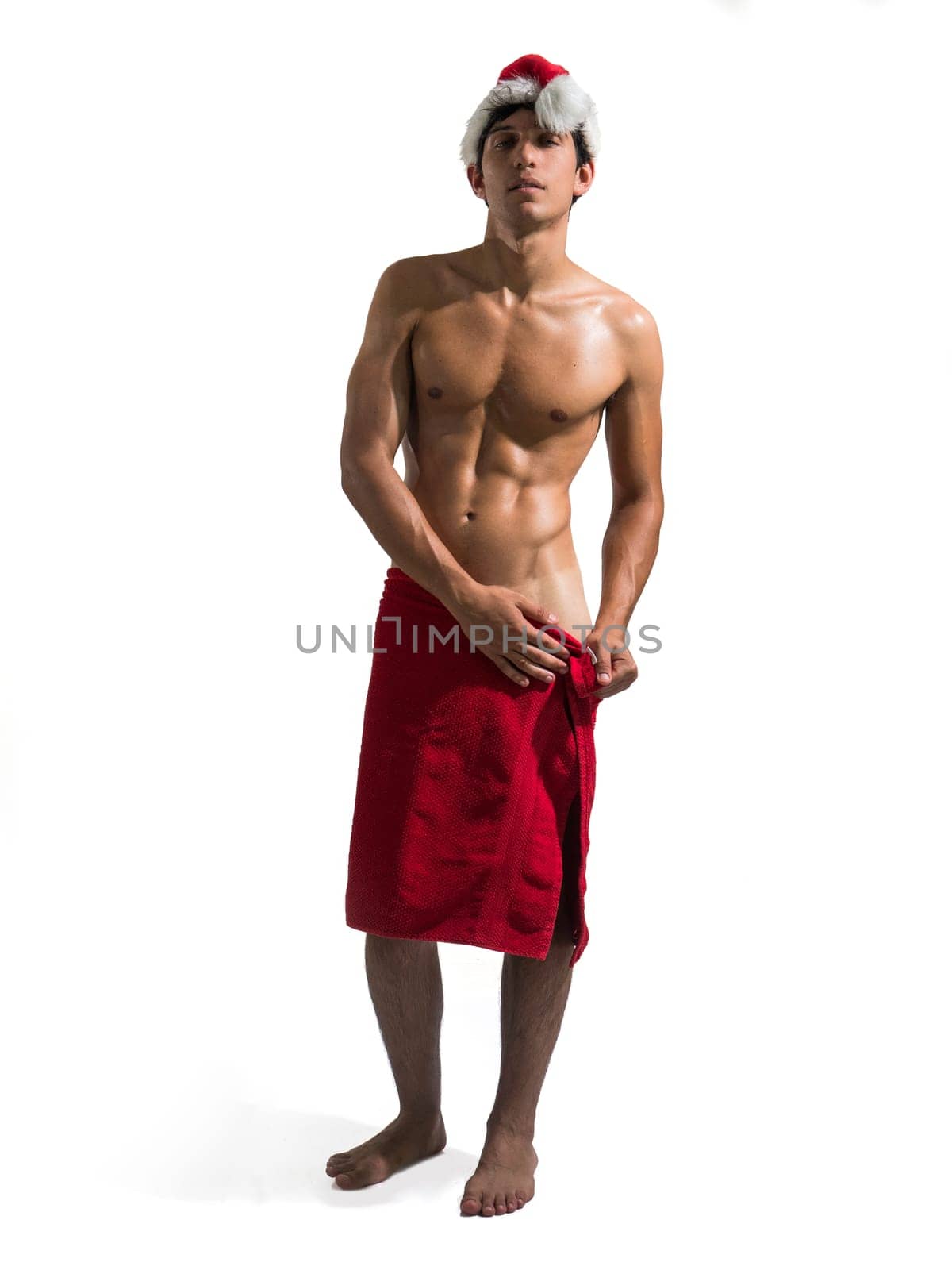Naked Young Attractive Fit Man Posing in Studio with White Background Covering Groin Area with Towel, Wearing Santa Claus Hat and Looking at Camera Smiling. Full figure shot