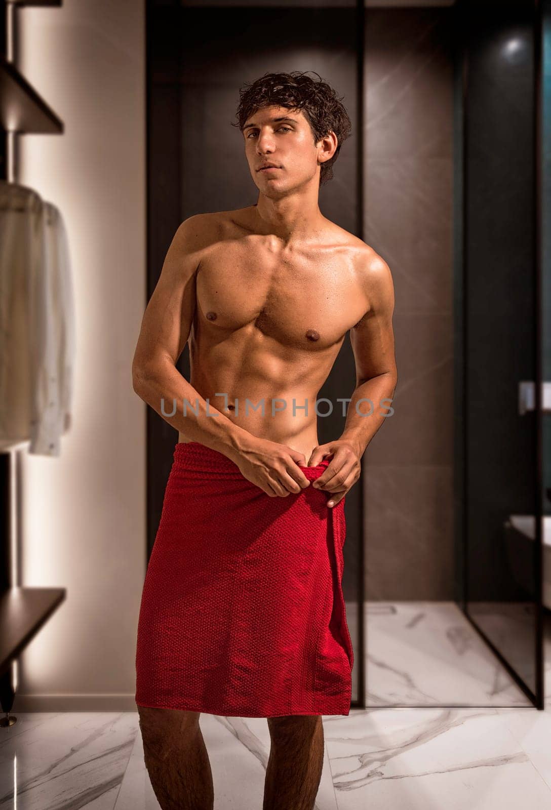 Handsome shirtless athletic fit young man going out of shower in room, with red towel wrapped around his waist