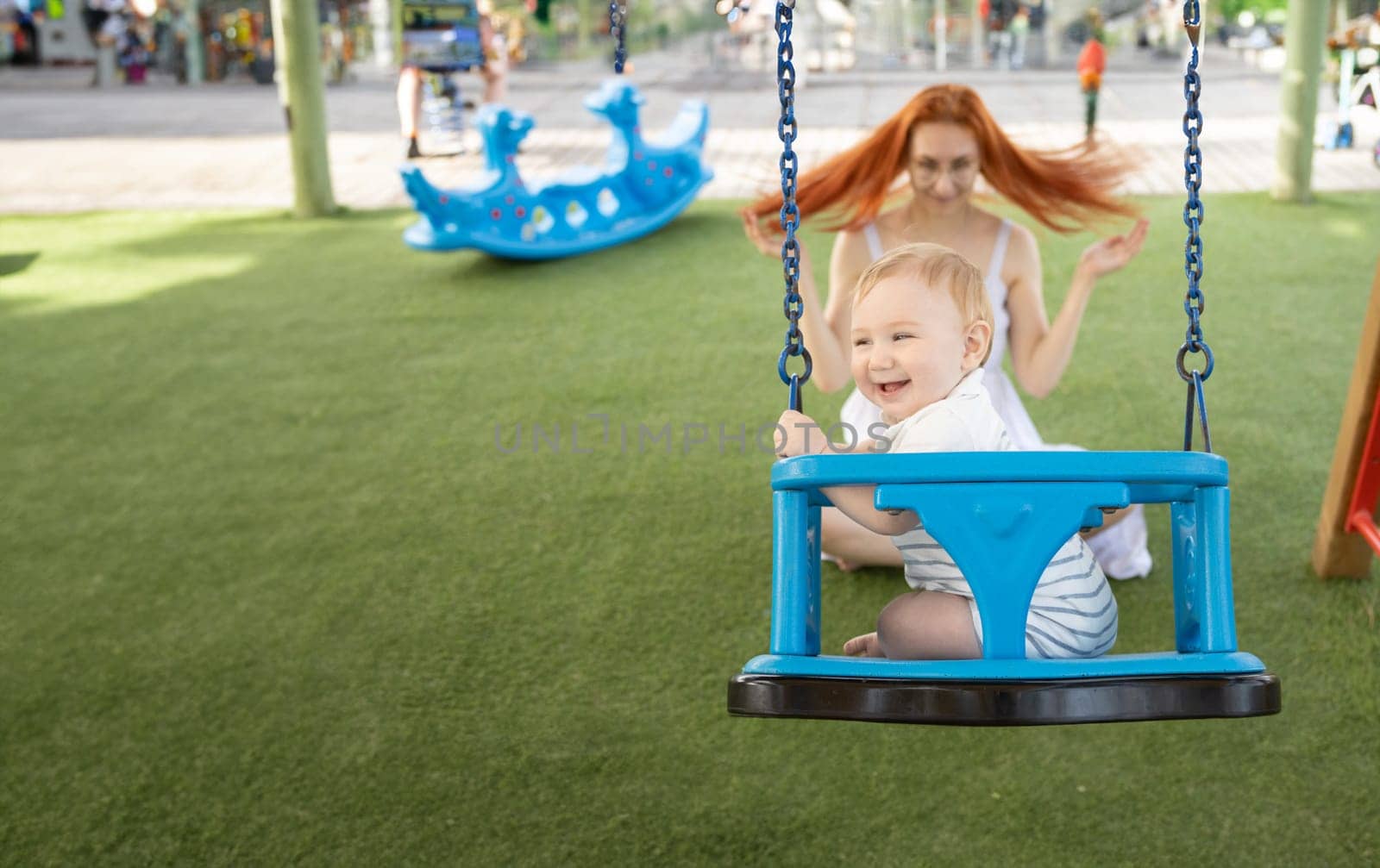 A little laughing boy on swings on an outside playground with his ginger mother by Studia72