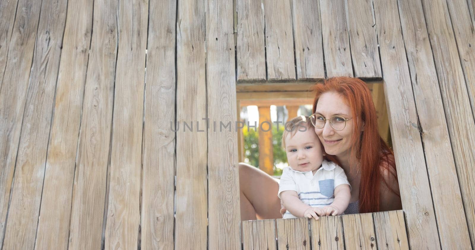 Cute baby and his mother sits in a wooden house on the playground - looking in the camera. Mid shot