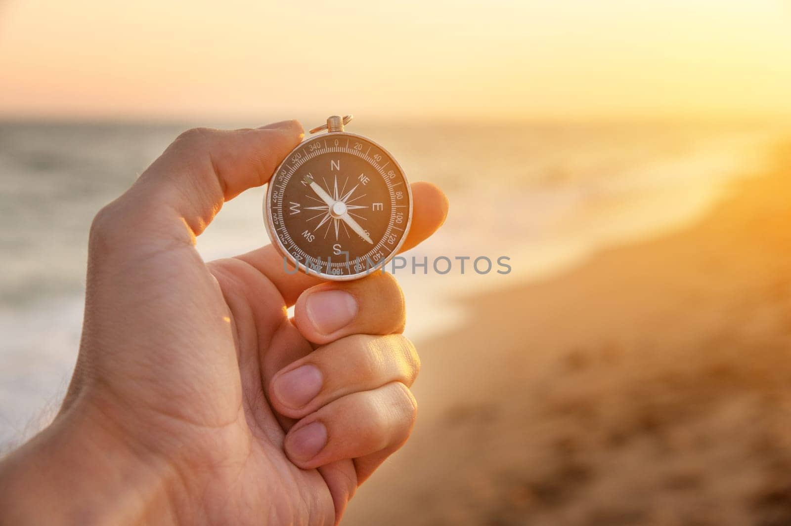 a man's hand holds a compass on the beach. Hand with compass over sand by the sea, direction and adventure concept
