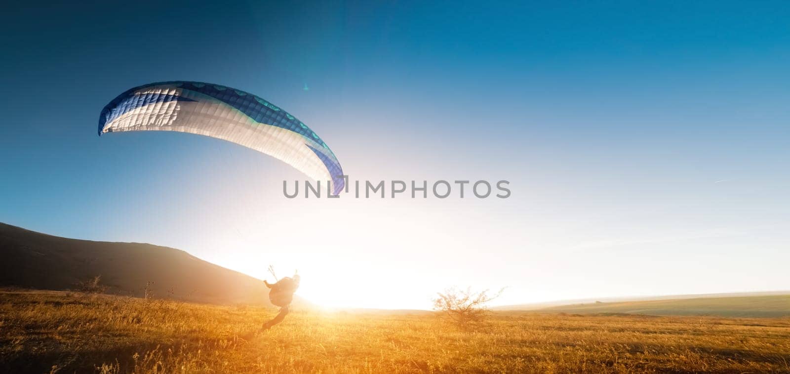 A paraglider glides along the ground at sunset with mountains in the background. Panoramic shot banner for paragliding in warm colors. Glare from the sun in the frame.