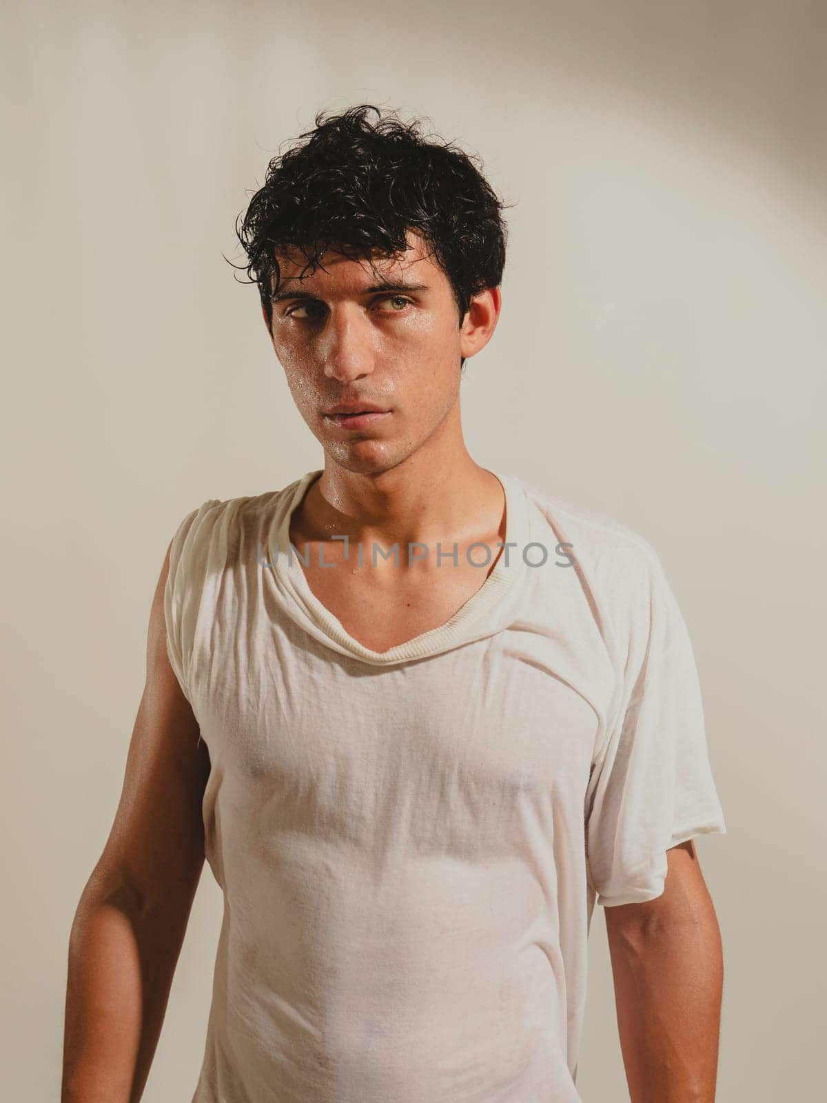 Serious looking young man wearing wet t-shirt, looking at camera on light background. by artofphoto