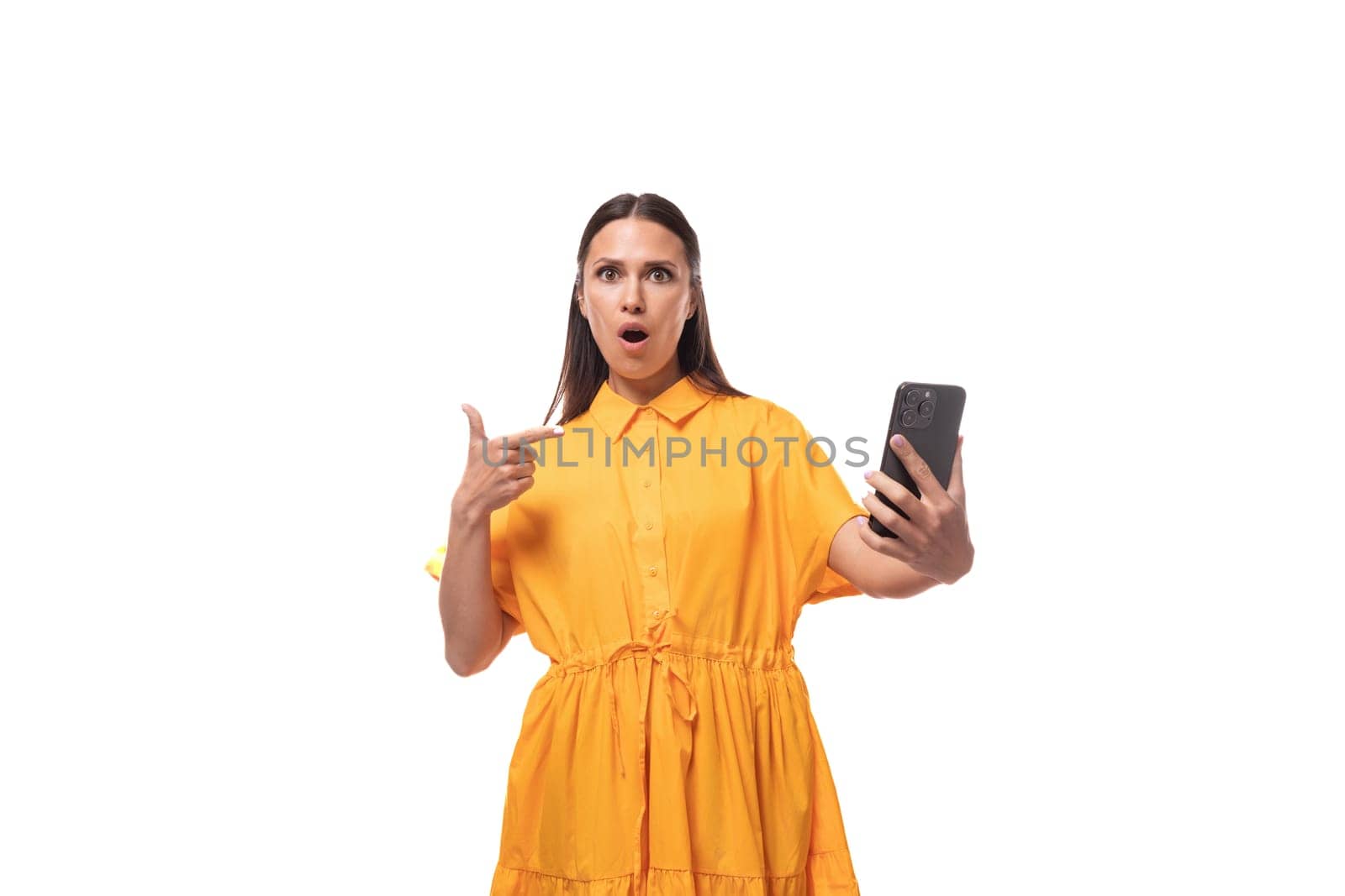 a young woman dressed in a summer yellow dress shows her hand in surprise at a smartphone on a white background by TRMK