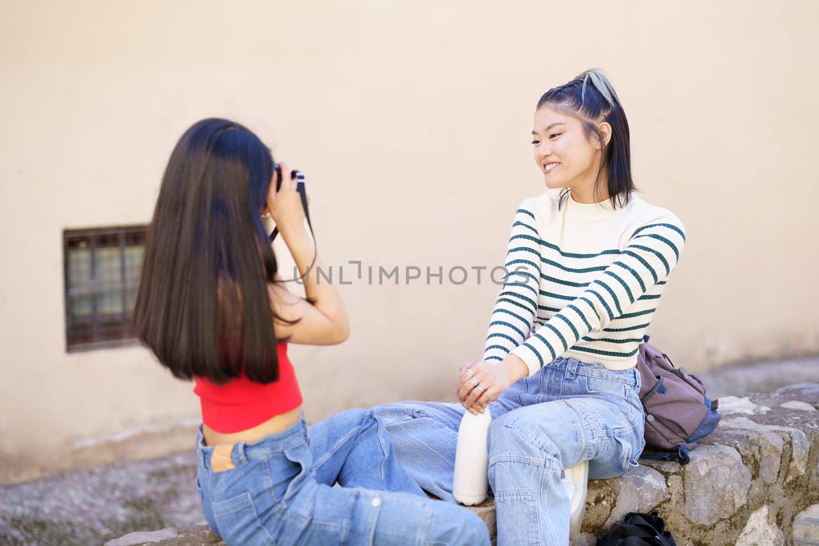 Young Chinese woman photographer taking photo of girlfriend showing peace gesture on city street during vacation