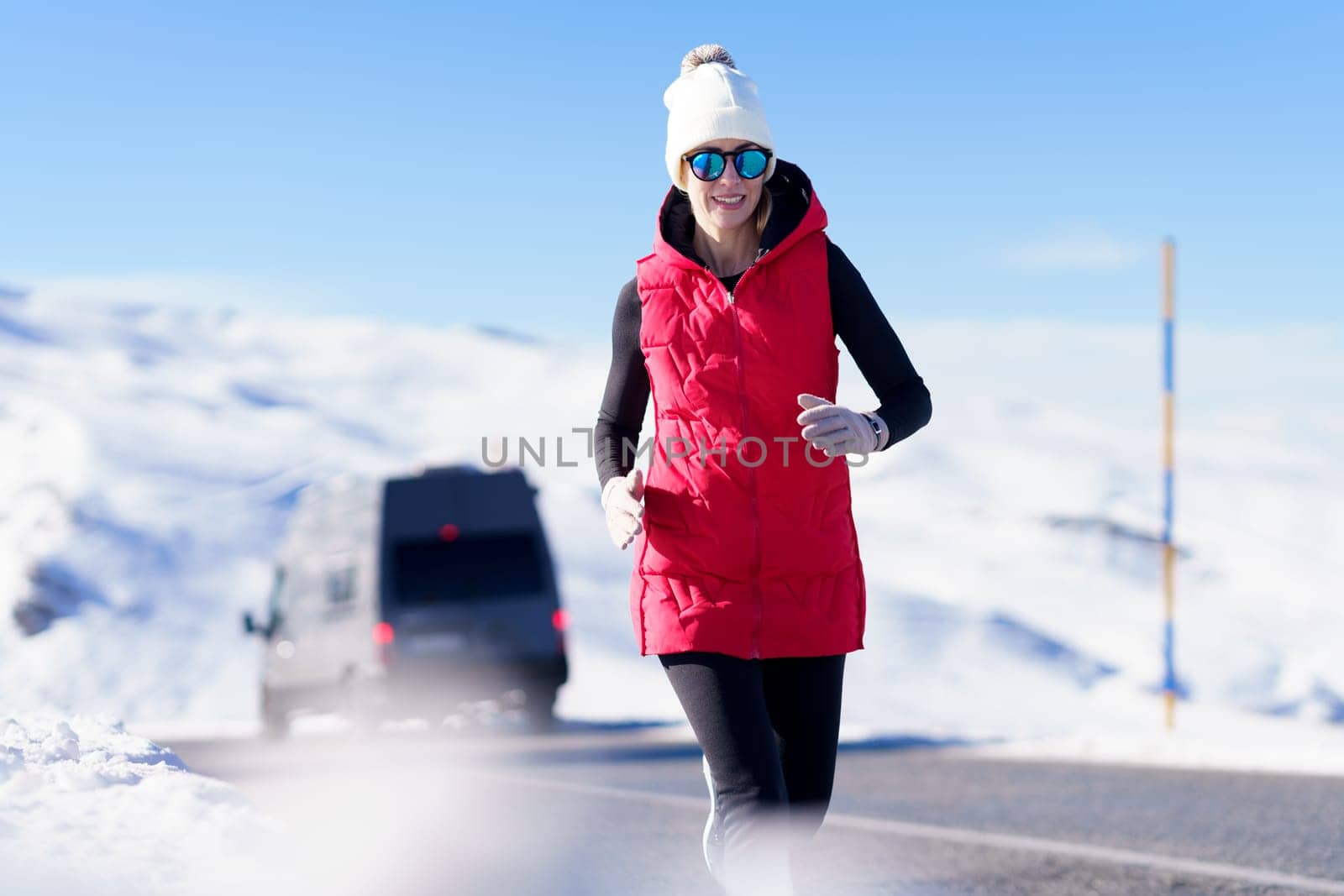 Smiling athletic woman in warm clothes and sunglasses running on roadway with snowy mountains on sunny day
