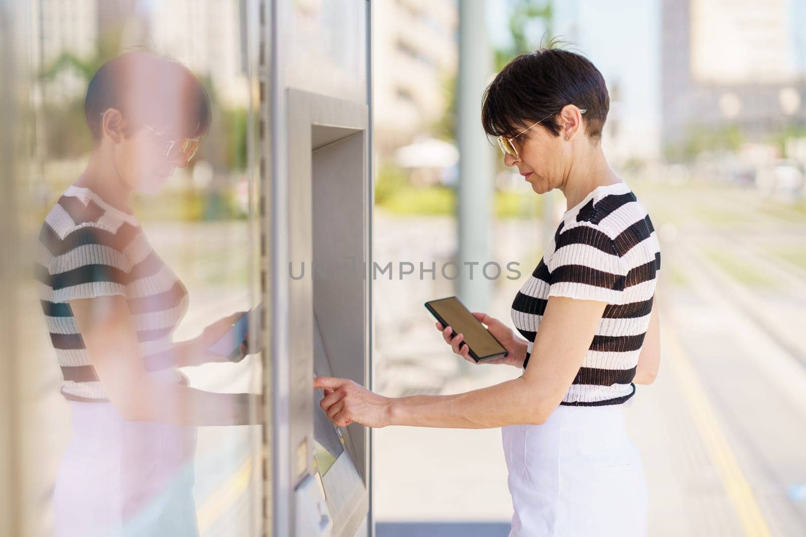 Concentrated adult female in casual outfit and sunglasses standing with smartphone and buying ticket on terminal on street