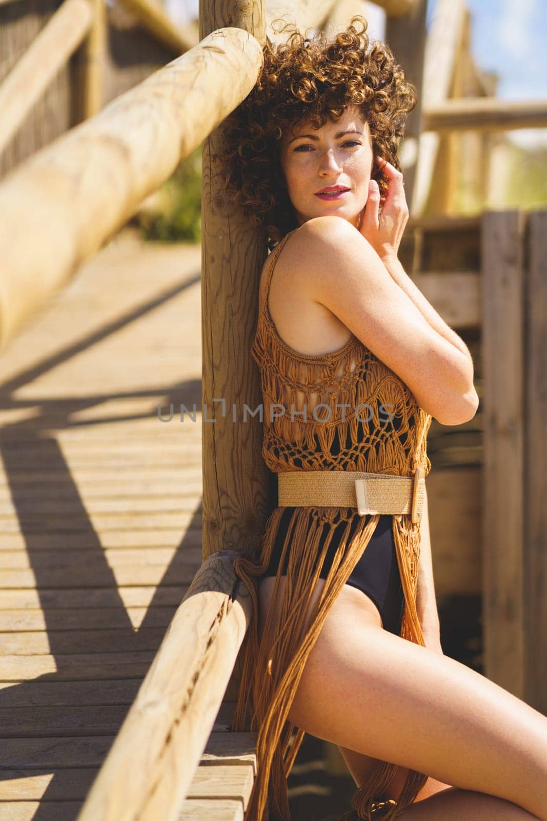 Young curly haired female wearing knitted beach cover up and swimsuit leaning on wooden railing touching face while looking at camera during summer day