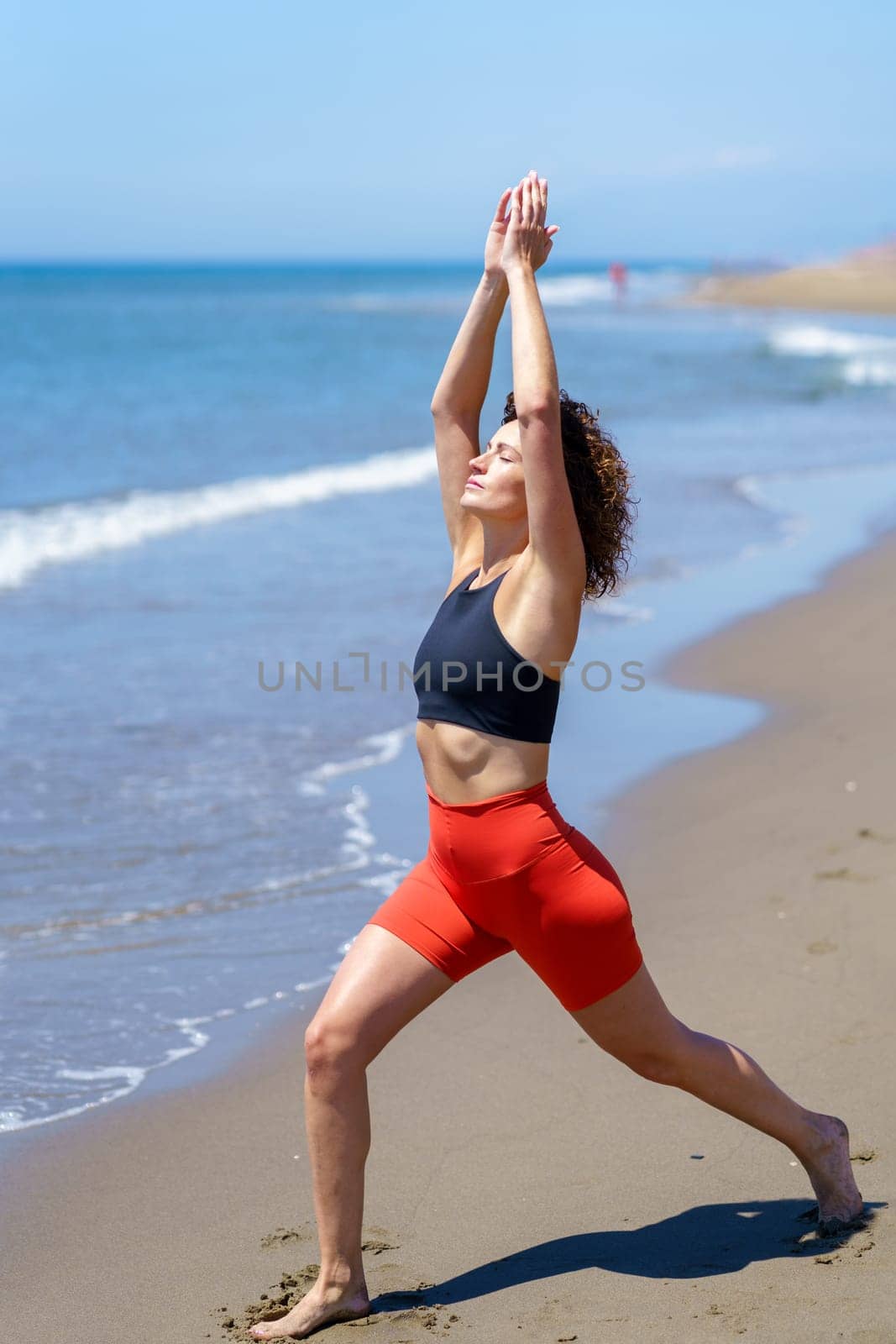 Full length of fit barefoot female with curly hair in sportswear doing Crescent Lunge pose while practicing yoga near waving sea