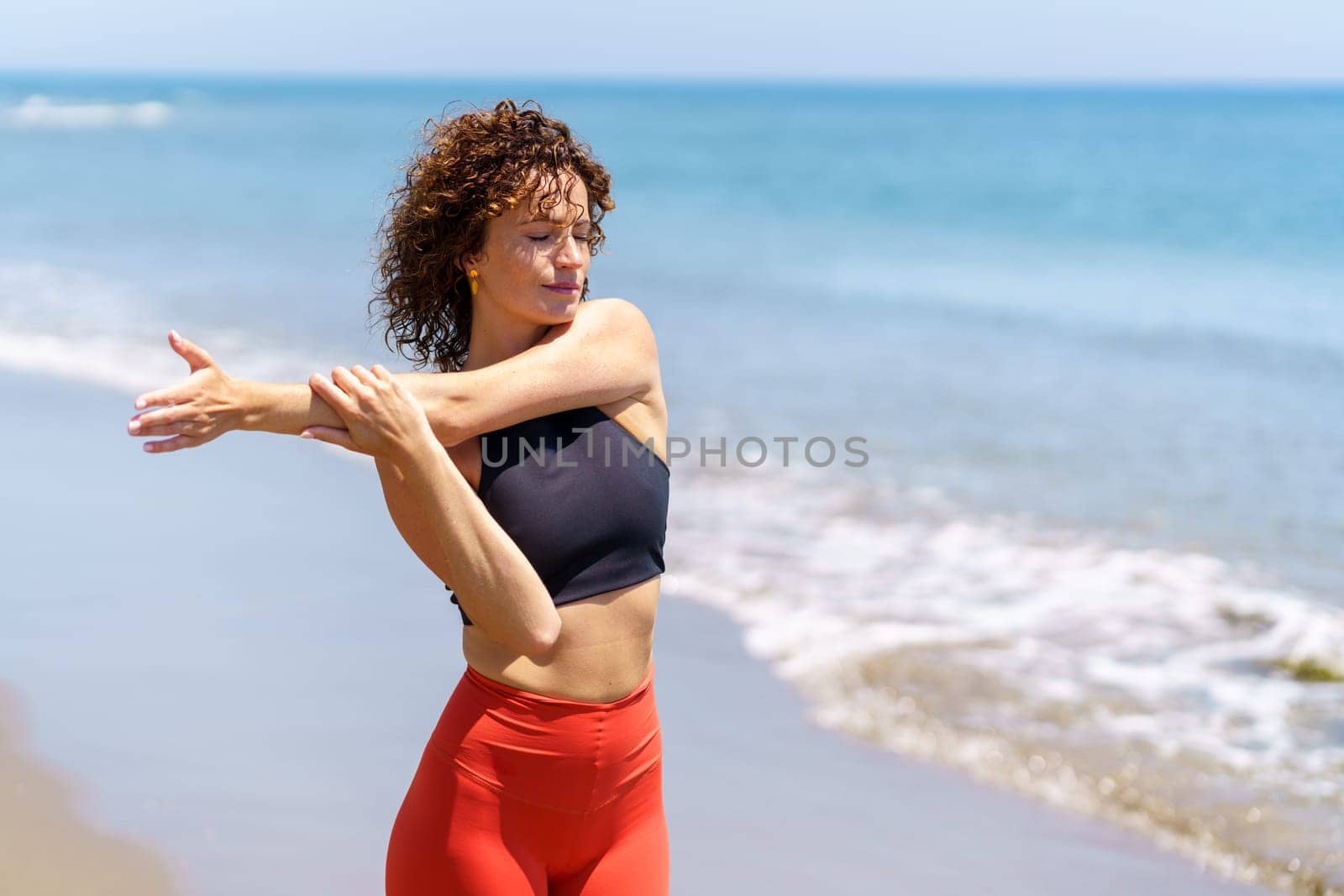 Pensive female athlete with closed eyes and curly hair stretching arm during workout on sunny day at beach