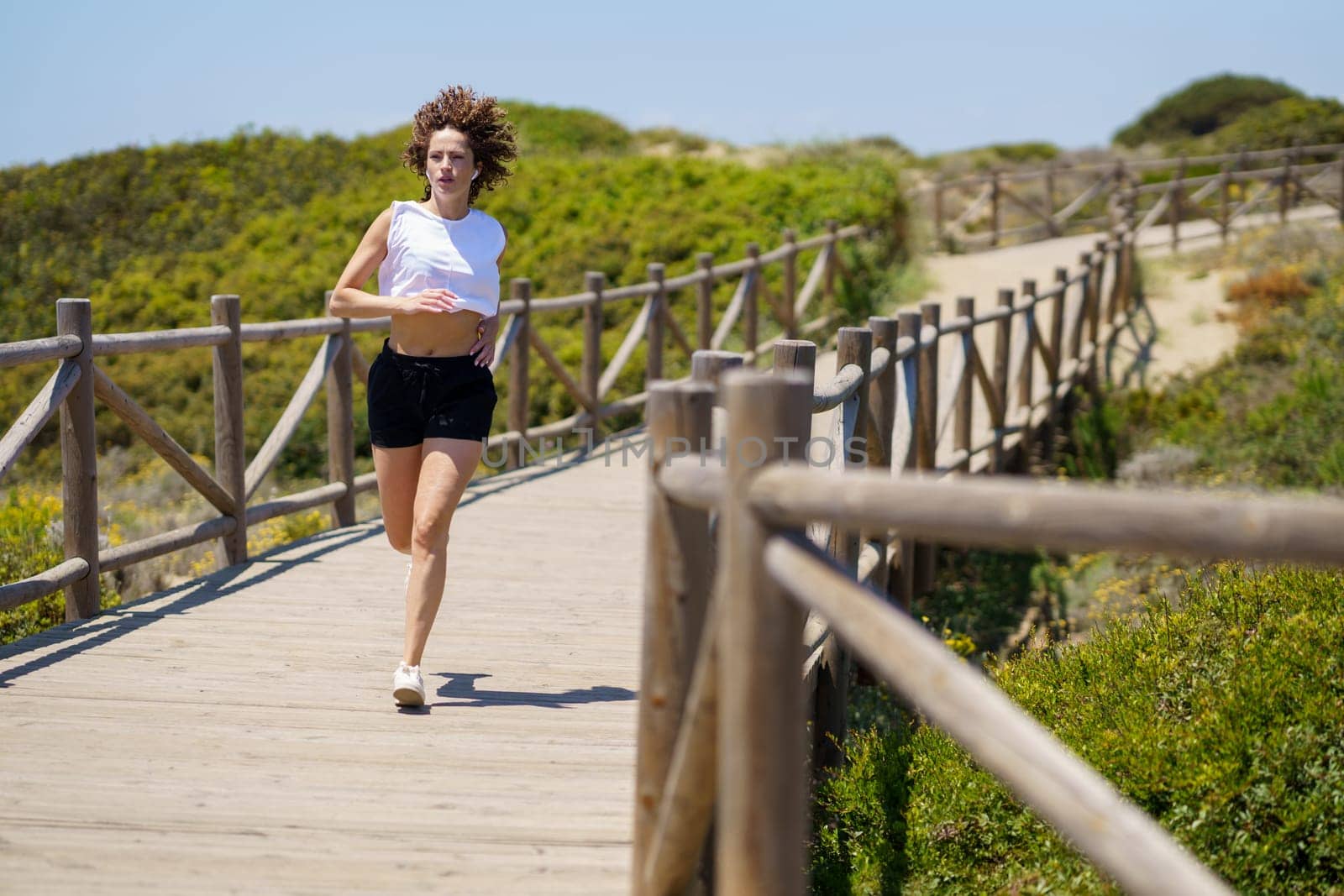 Full body of young active female in sportswear jogging on wooden walkway during fitness workout on sunny summer day