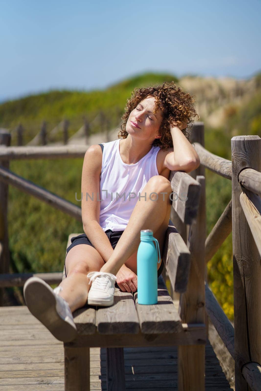 Smiling young female with curly hair eyes closed, relaxing on wooden bench and leaning head on hand while sitting on wooden bench with water bottle on bridge