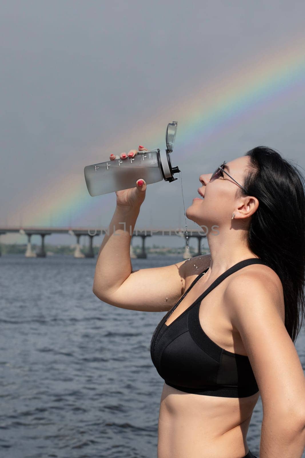 A beautiful and slender girl in sunglasses pours water over herself from a sports bottle after a fitness workout on the street in the summer, against the sky with a rainbow. Sport concept. Close-up.