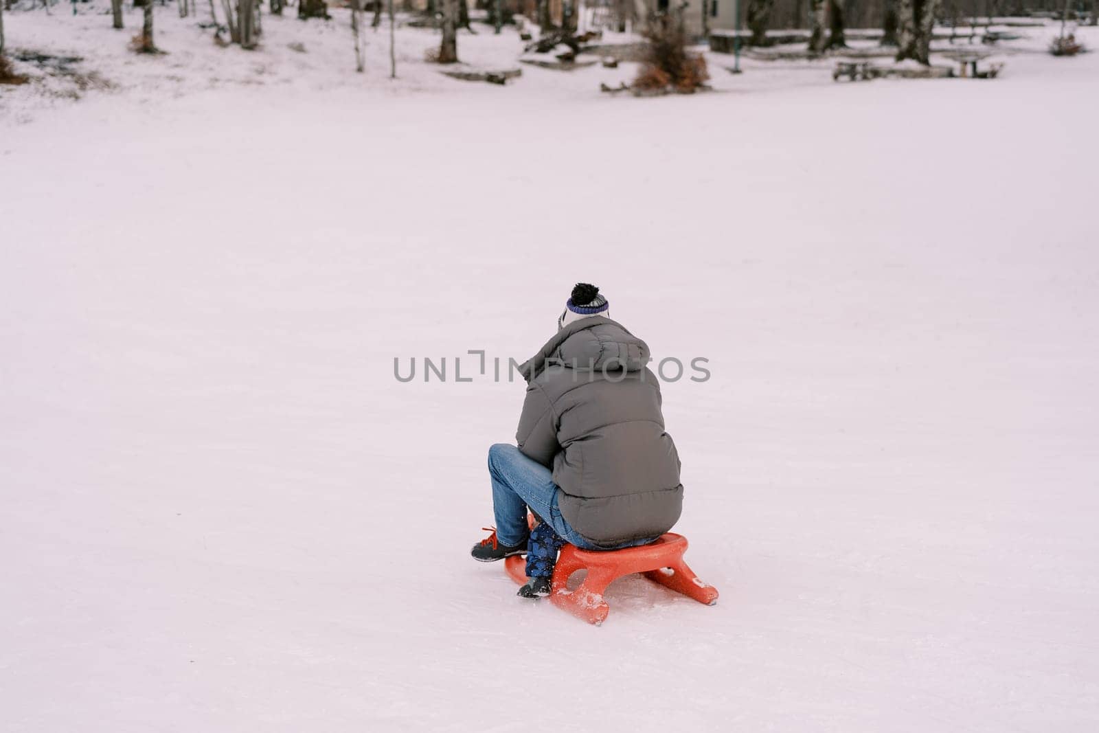 Dad and a small child are sledding on a snowy hill. Back view by Nadtochiy