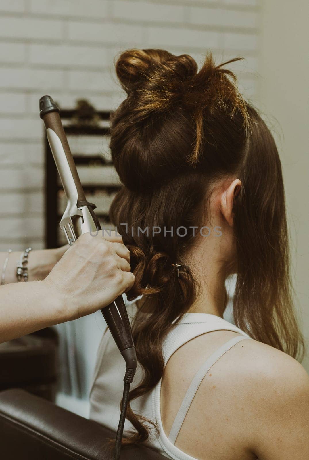Portrait of a young Caucasian girl sitting from the back in a hairdresser's shop, where the master is curling her hair on a flat iron, close-up view.