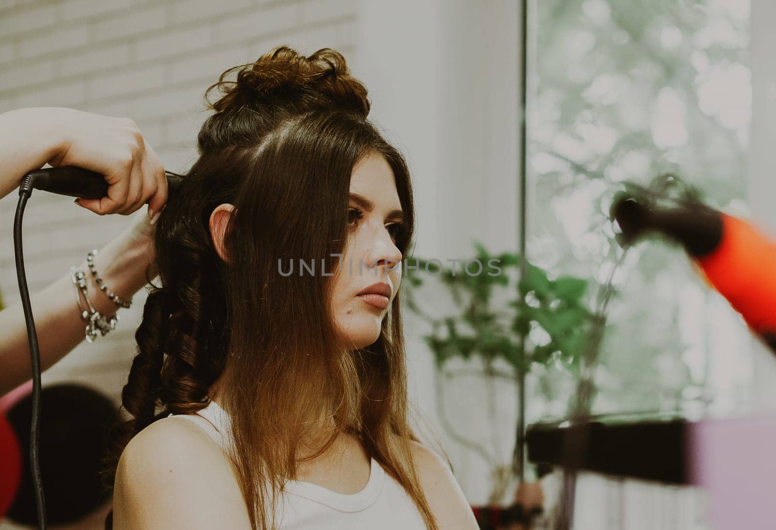 Portrait of a young beautiful brunette Caucasian girl sitting in a hairdresser's shop, where the master is curling her long hair on a flat iron, close-up view from below.