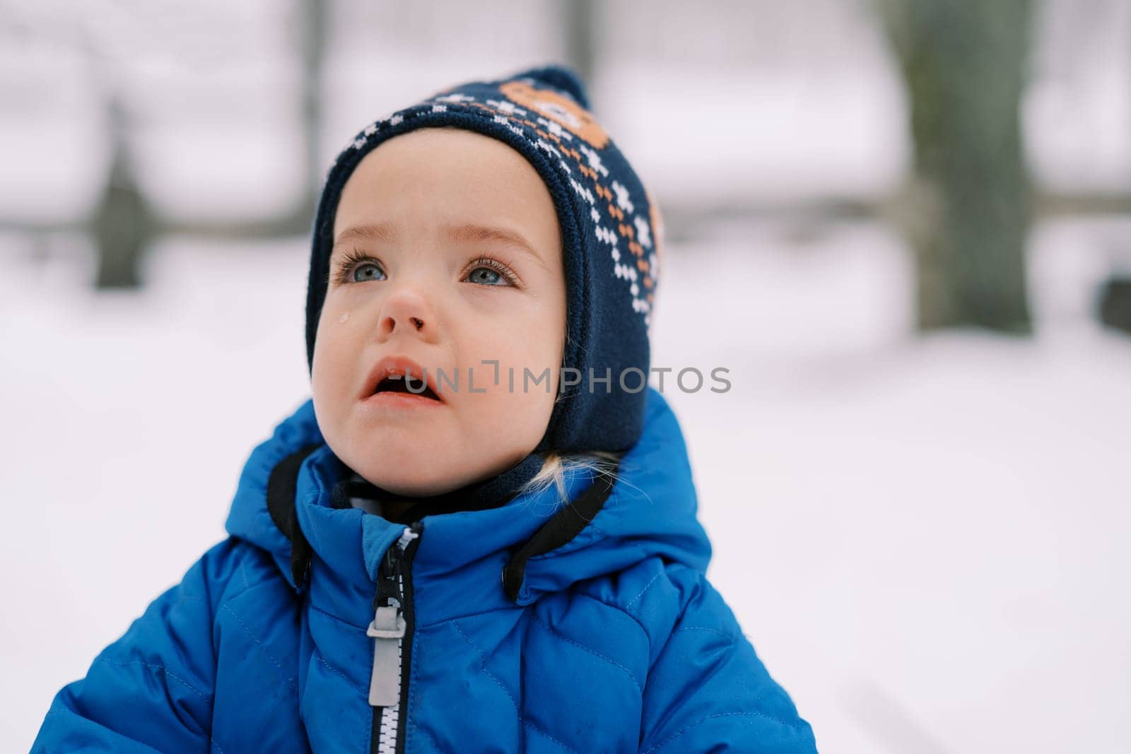 Little crying girl with tears in her eyes stands in the snow and looks up by Nadtochiy