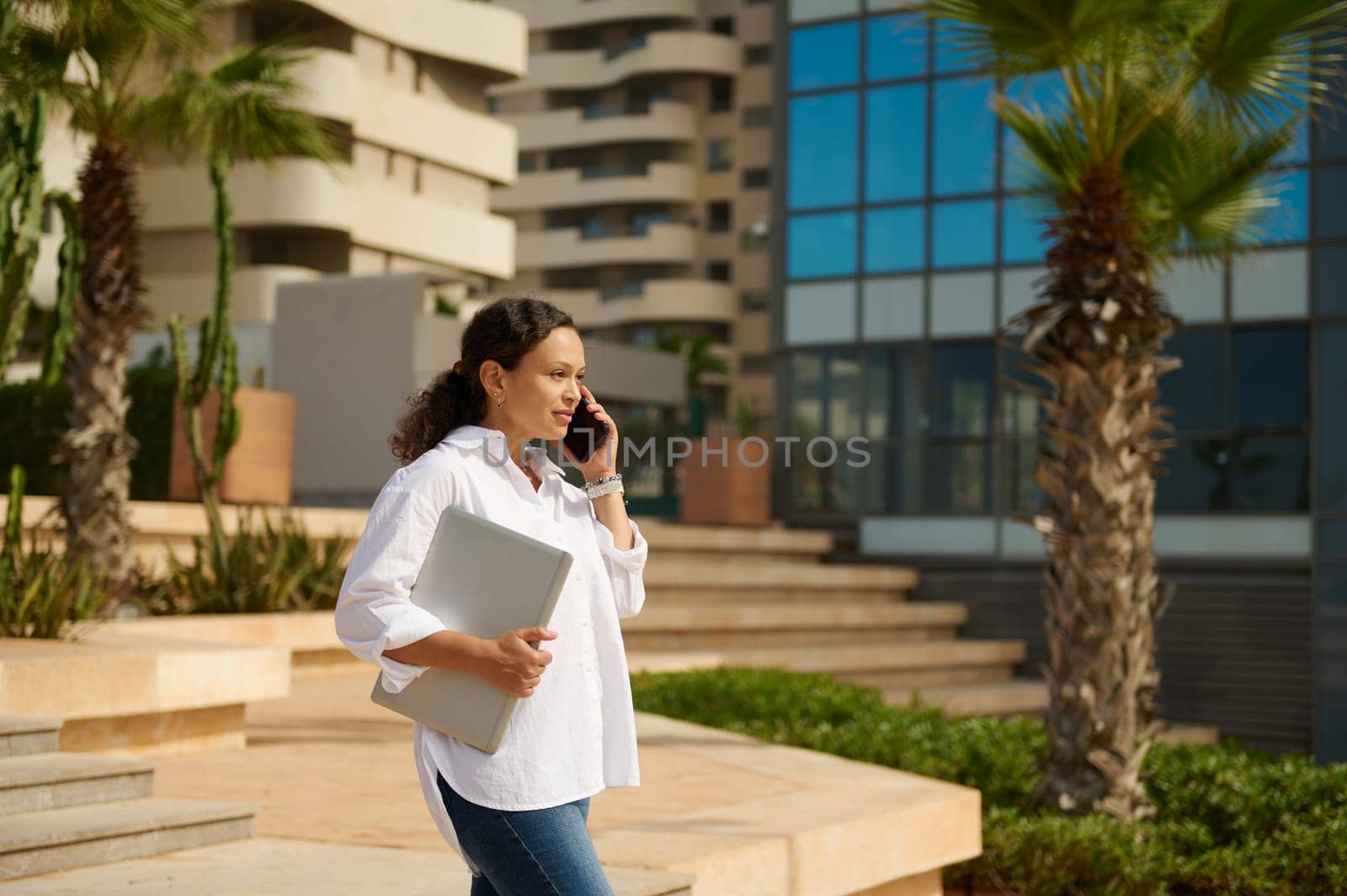 Confident multitasking woman entrepreneur talks on mobile phone, holds laptop, walks on the the urban street against modern high rise buildings background. People. Business. Real Estate investment