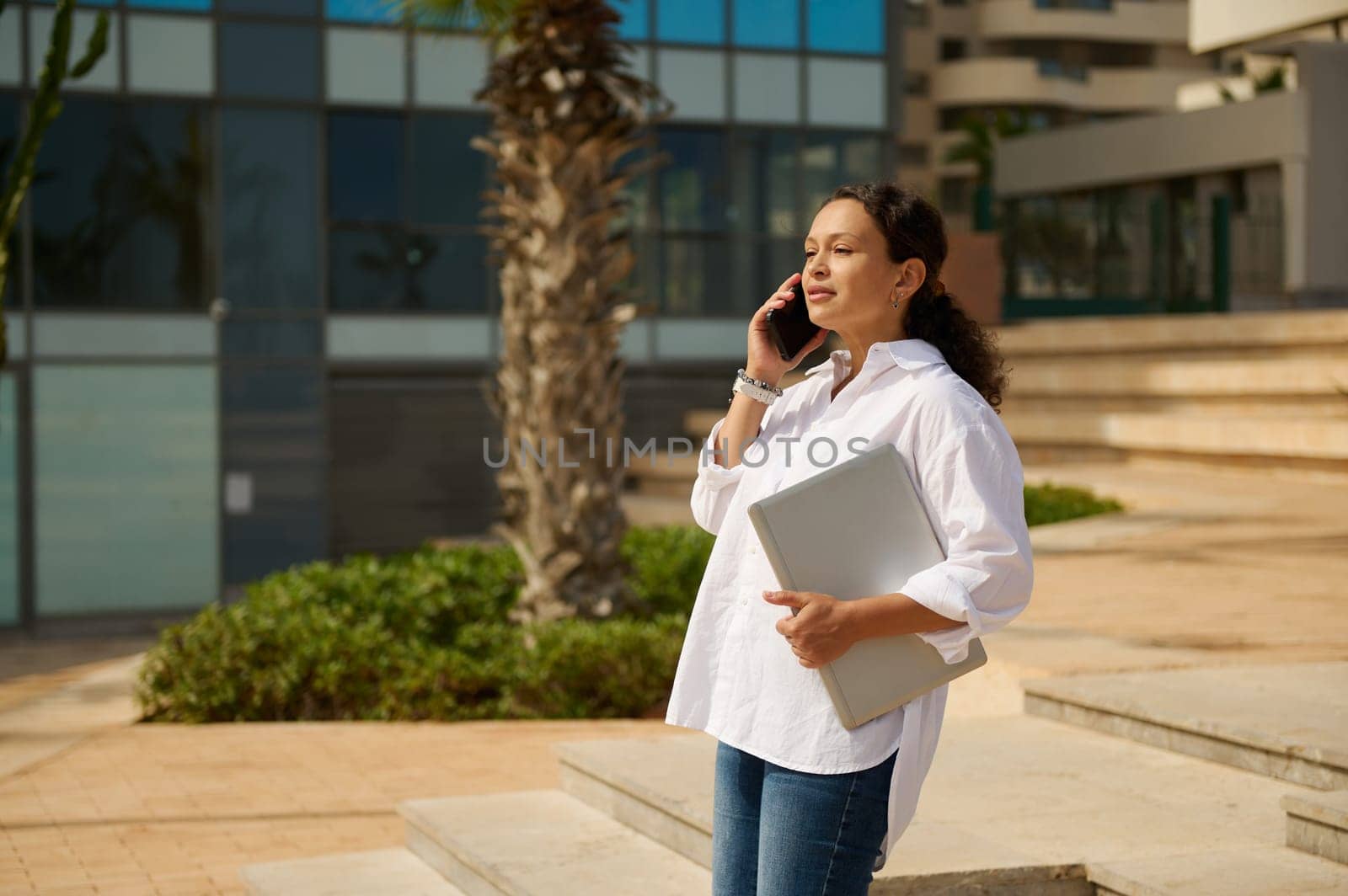 Beautiful Latin American young woman, real estate agent, freelancer, sales manager standing against cityscape background and talking on cellphone. People. Business. Recruitment. Real Estate investment