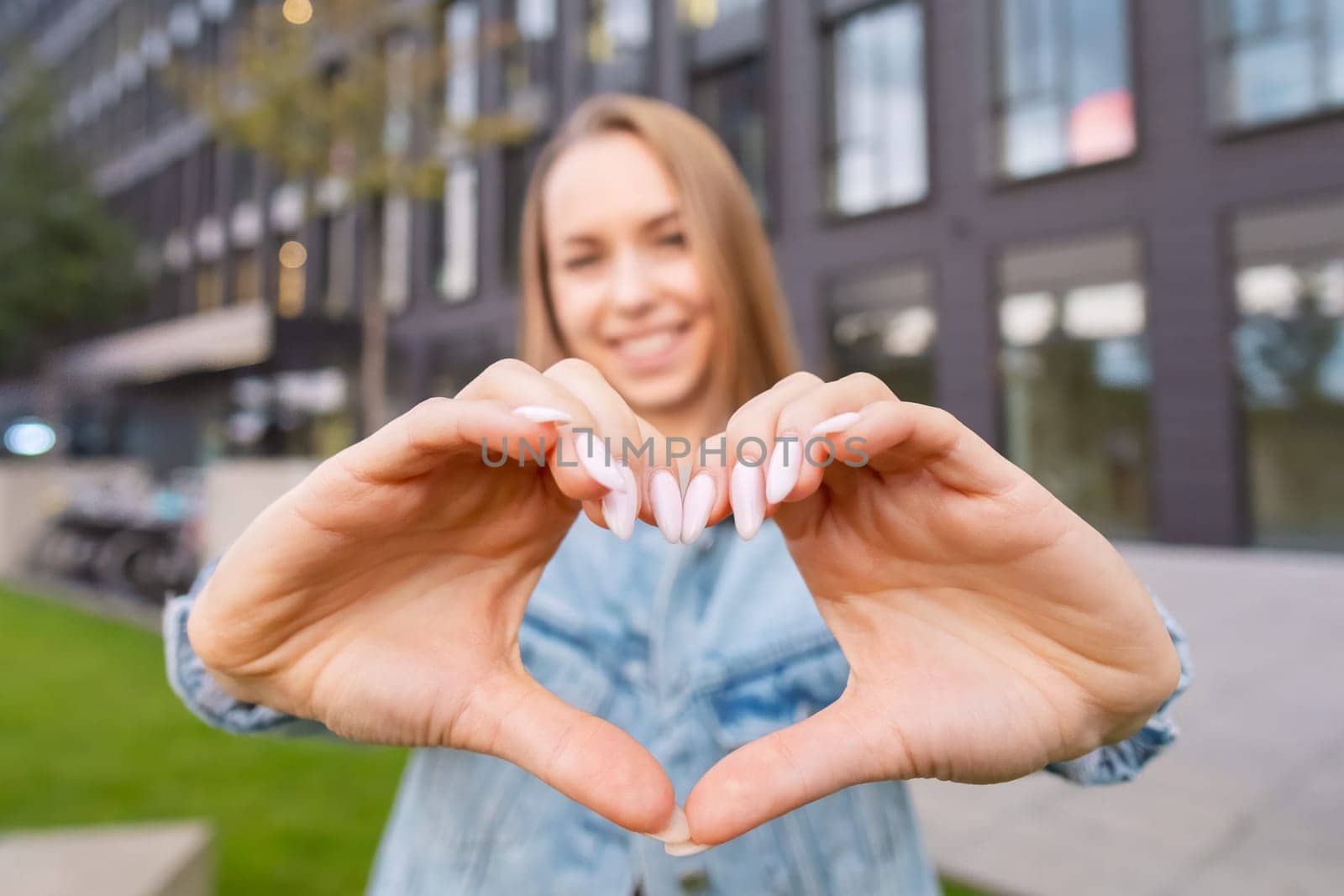 A stylish blonde woman in a casual jean jacket stands near a contemporary building, expressing her affection by forming a heart shape with her hands.