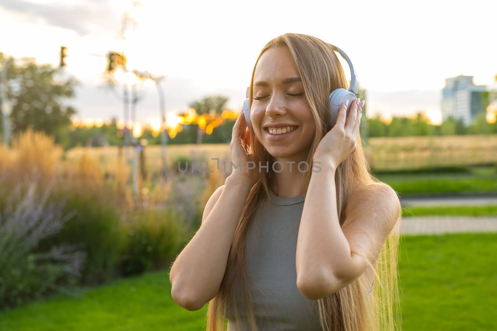 Cheerful young woman enjoys listening to music and singing while sitting in park. Happy lady in headphones rests spending time in nature