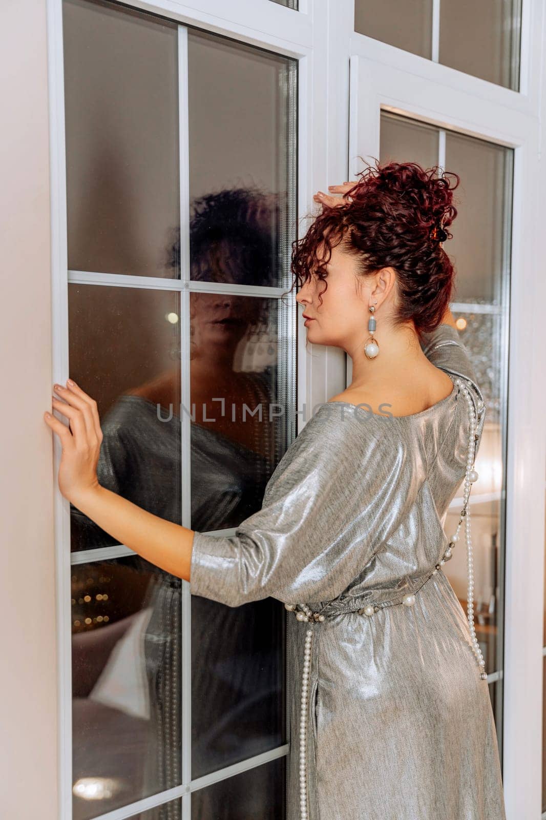 Woman portrait window. Curly-haired middle-aged woman looks out the dark window at her reflection. She is dressed in a silver dress with pearl beads and earrings.