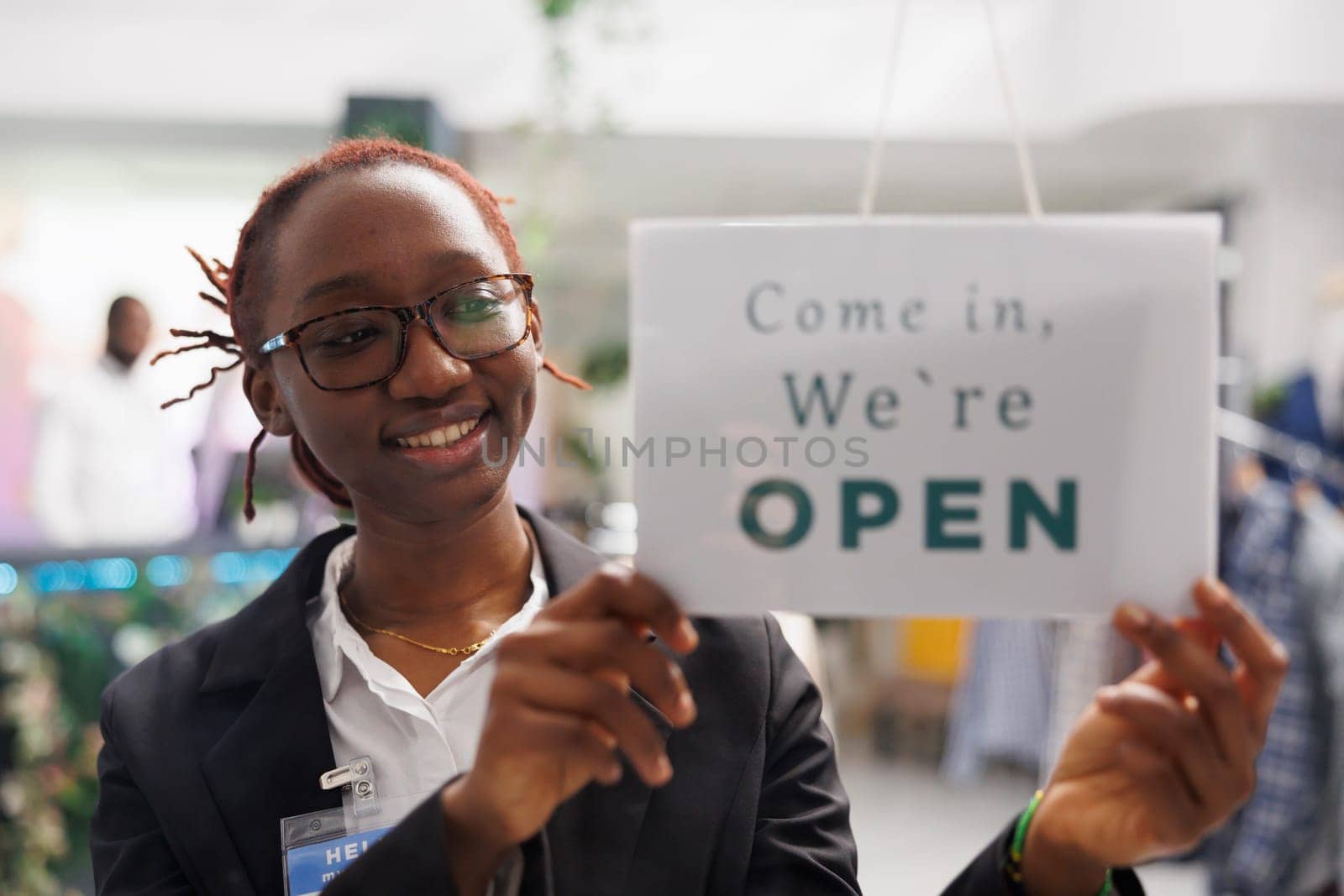 Clothing store smiling african american woman assistant hanging opening signboard on entrance door. Cheerful fashion shop worker putting sign on window noticing about workday beginning