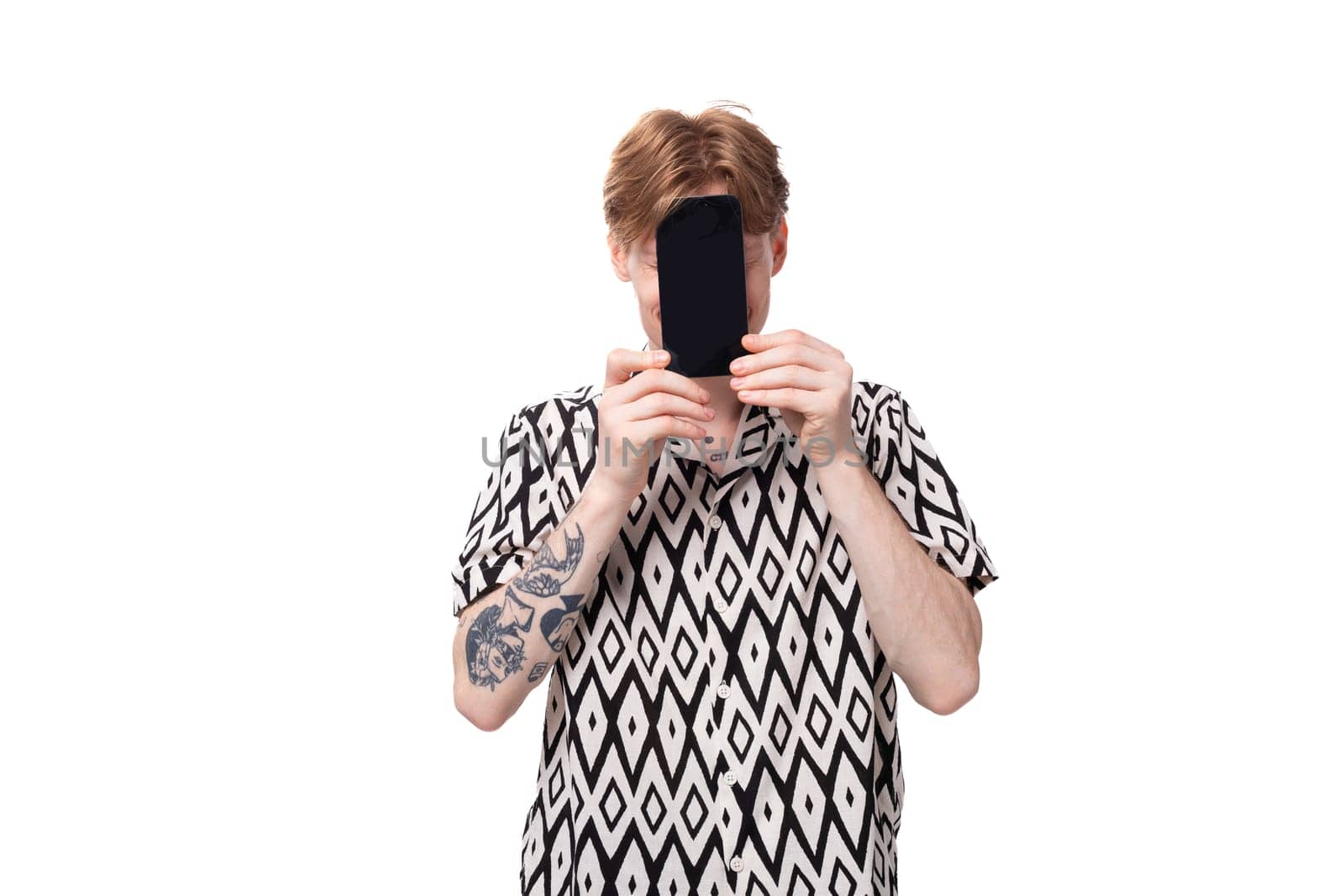 young smiling red-haired man dressed in a summer short-sleeved shirt in black and white holds a smartphone with a mockup.