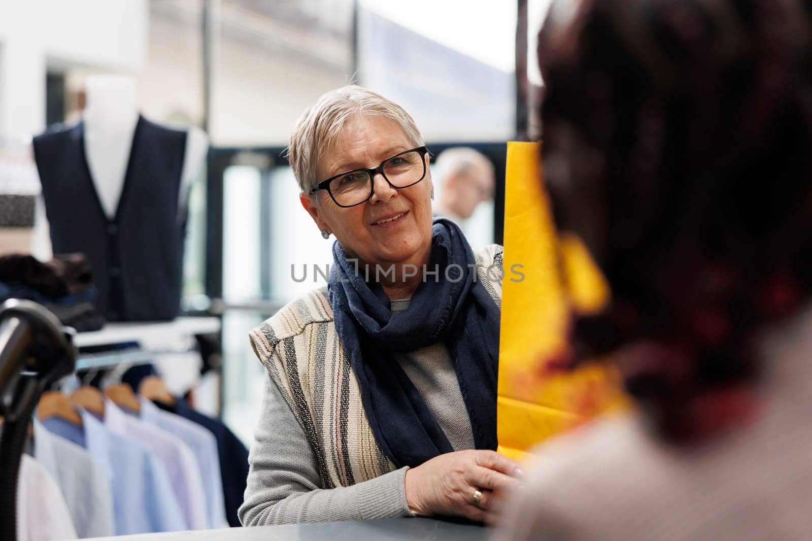 Elderly shopper taking shopping bags from employee, making purchase at counter desk in clothing store. Senior woman buying fashionable formal wear in modern boutique. Commercial activity concept