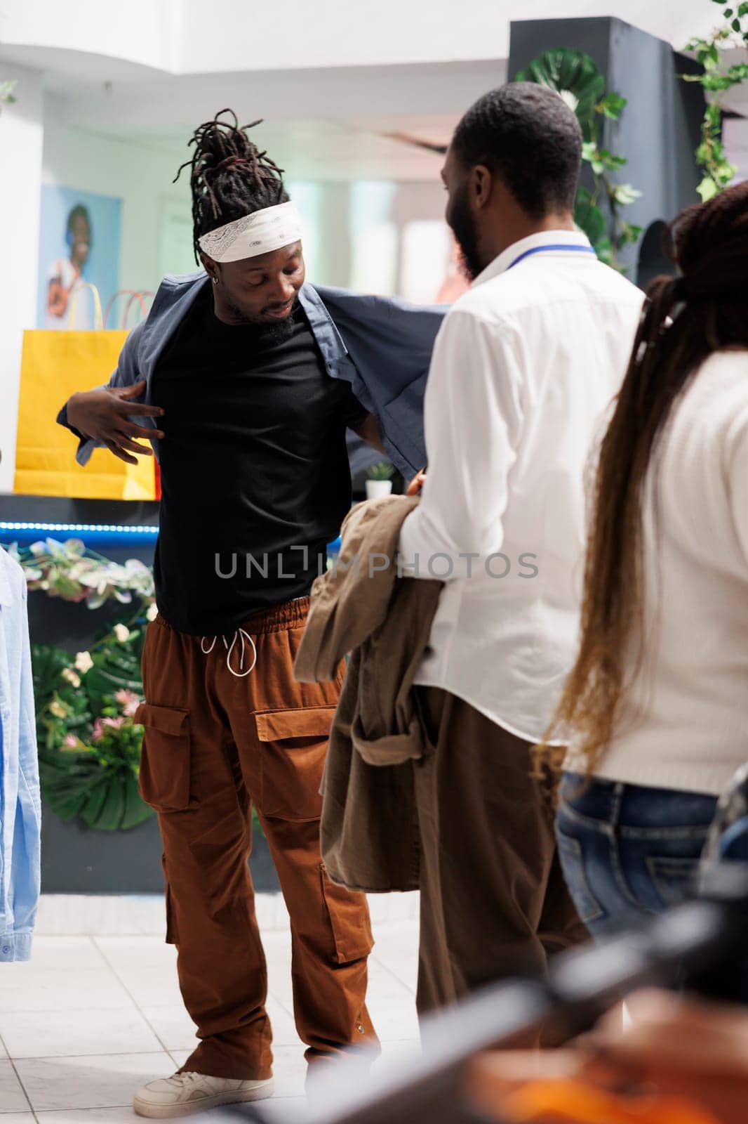 Shopping mall assistant helping customer to try clothes before buying in fashion showroom. Young african american man wearing shirt while checking apparel size in modern store