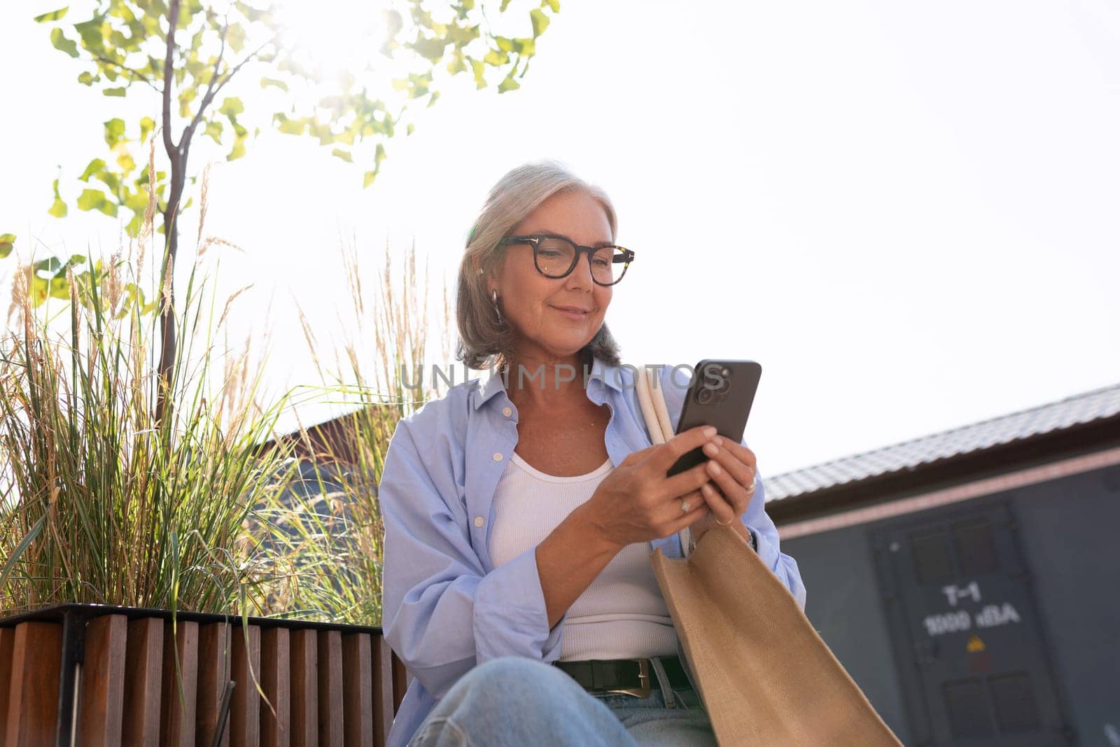 a pretty fashionable middle-aged woman with gray hair dressed in a summer style sits on a bench in the city and uses a gadget.