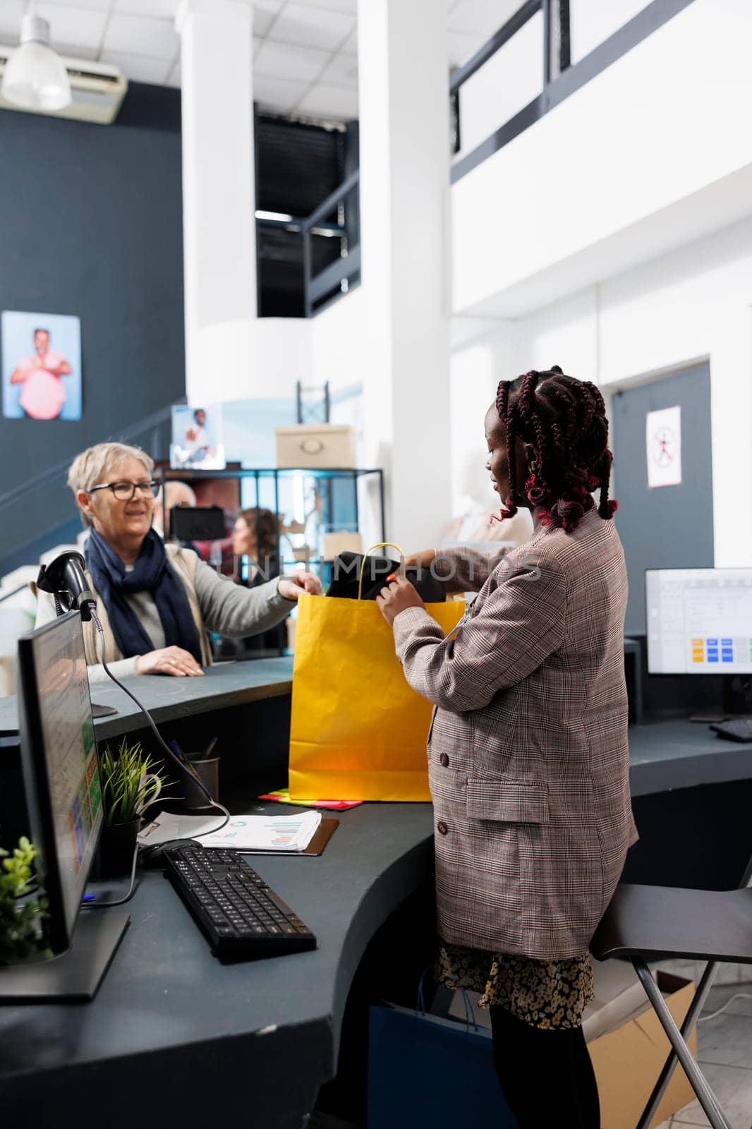 African american employee preparing bag with purchase for elderly client in modern boutique. Fashionable woman buying stylish casual wear in store, making electronic payment at pos terminal.