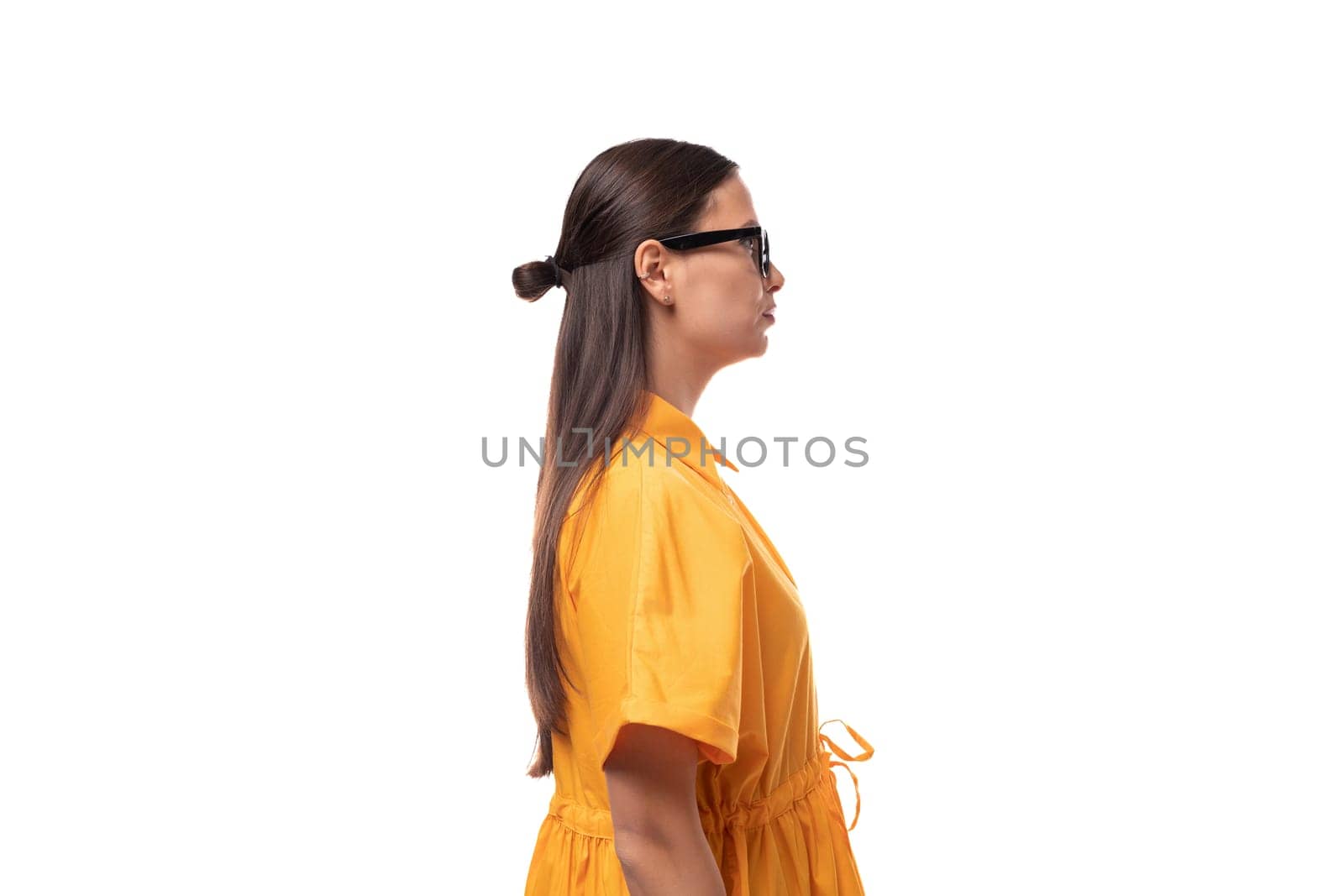 European young woman with black hair dressed in an orange summer dress stands sideways.